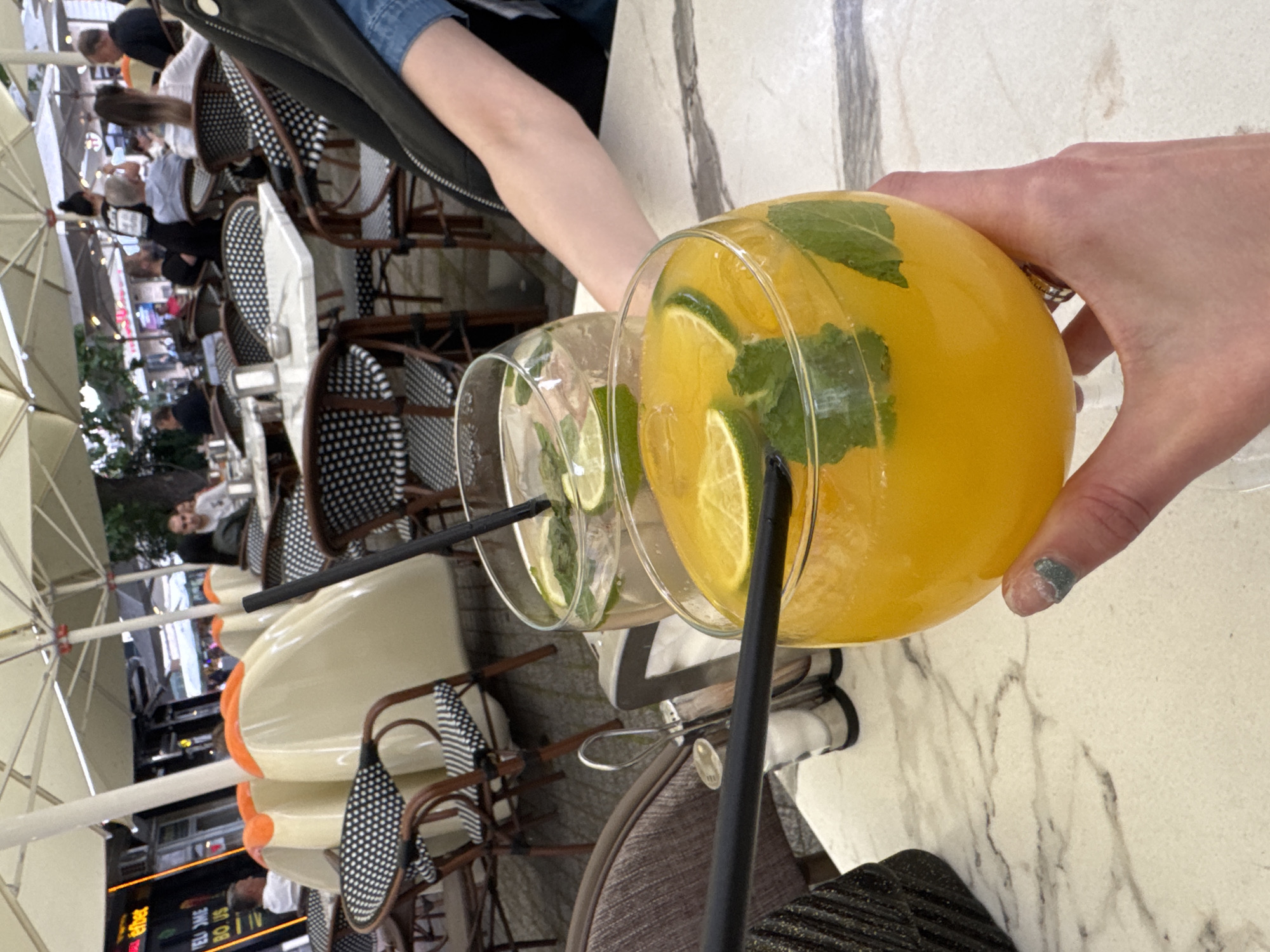 The image shows two cocktails on a table in what appears to be an outdoor cafe setting. One cocktail is bright orange in color, contains lime slices and mint, and is being held by a person's hand. The other cocktail is lighter, likely clear, and also contains lime slices and mint. Both have black straws. The background reveals tables and chairs, suggesting a busy cafe environment, possibly in Plovdiv, Bulgaria, given the presence of umbrellas and people enjoying the outdoor setting. The chairs have black and white patterns. A street scene with buildings is visible in the background.