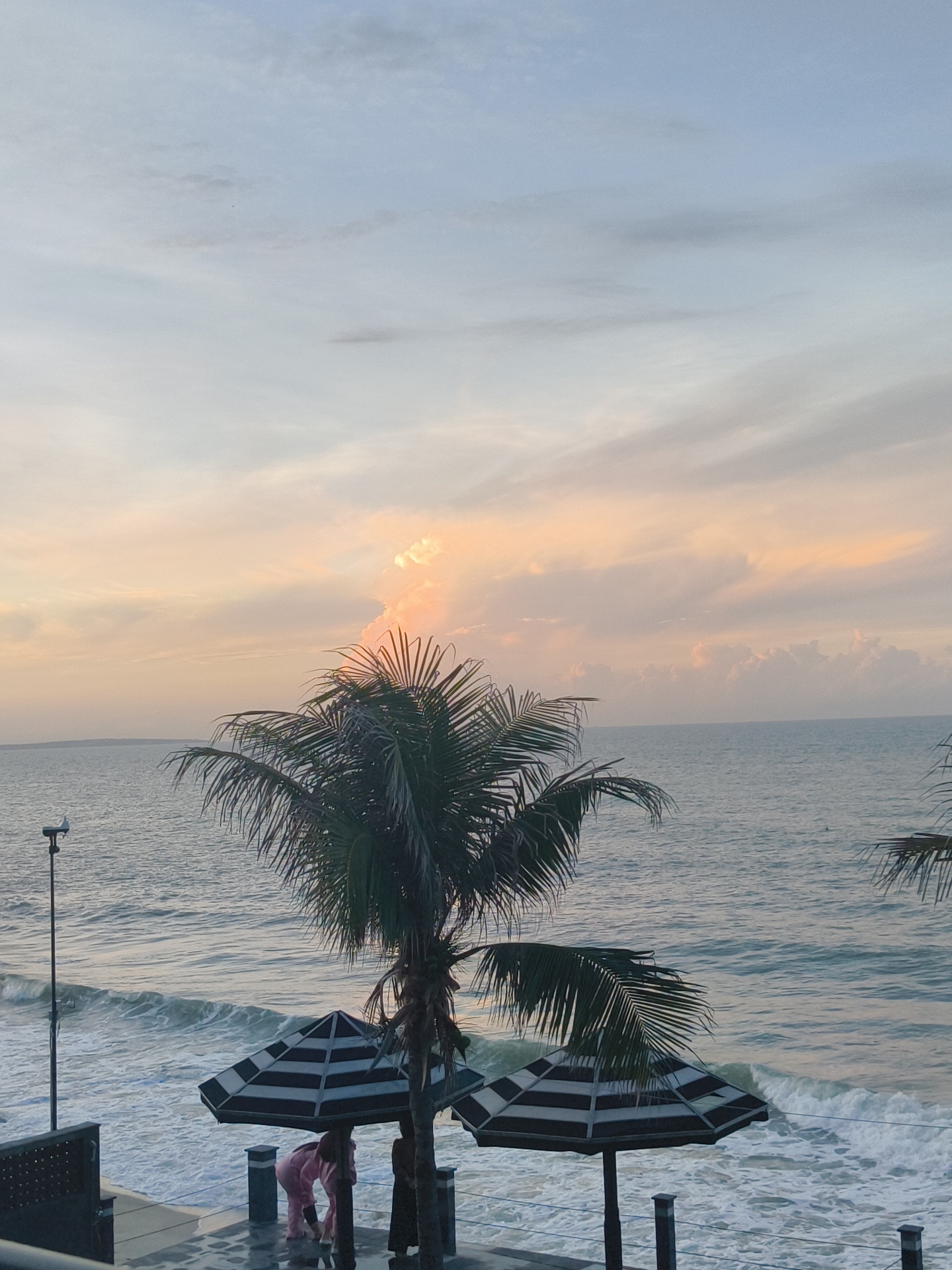 A low-angle shot captures a serene beach scene at dusk in Phu Thuy Ward, Vietnam. The sky transitions from a pale blue at the top to hues of orange and pink near the horizon, with wispy clouds illuminated by the setting sun. The ocean, a calm expanse of blue-gray water, gently laps against a sandy shore, with white foam indicating breaking waves. In the foreground, a tall palm tree with lush green fronds stands prominently, its leaves rustling in the breeze. Two striped beach umbrellas, black and white, are positioned beneath the palm tree. Two individuals, one wearing a pink outfit, are interacting near the umbrellas. A thin lamppost with a bird perched on top is visible to the left of the palm tree. The scene is framed by a dark railing in the lower left corner.