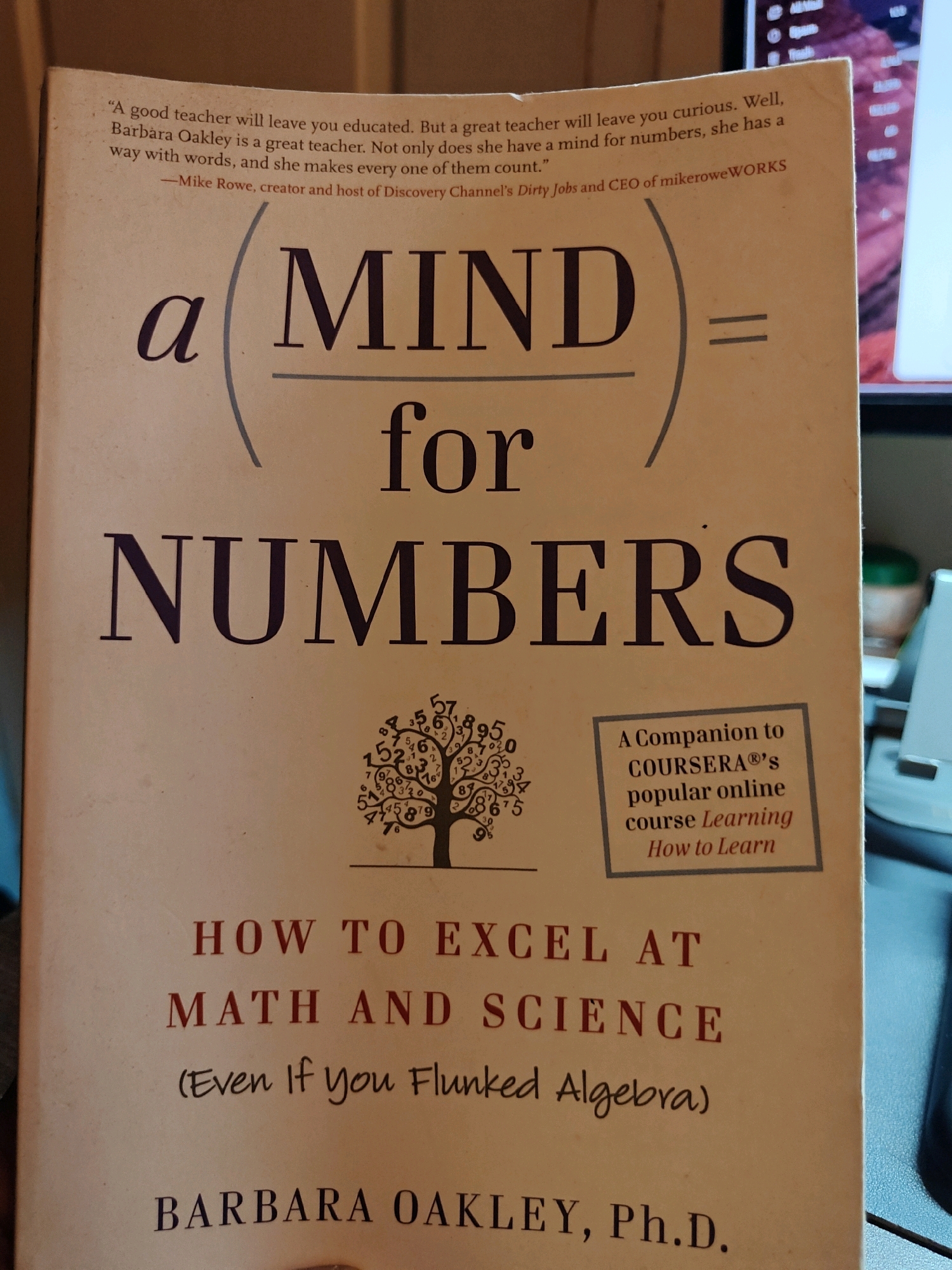 The image shows the cover of a book titled "A MIND for NUMBERS" by Barbara Oakley, Ph.D. The cover is primarily light beige and features a large, stylized equation: "a MIND / for NUMBERS =". Below this, in smaller red lettering, is the subtitle "HOW TO EXCEL AT MATH AND SCIENCE (Even If You Flunked Algebra)". A decorative tree made of numbers is visible on the right side of the cover. In the lower part of the cover, the author's name, BARBARA OAKLEY, Ph.D., is printed in dark red. To the right of the numbered tree, there is a grey box with text that reads "A Companion to COURSERA's popular online course Learning How to Learn". The lighting suggests an indoor environment, likely a room with artificial light. There are no people visible in the image. The image is taken from a slightly downward angle, focusing on the book cover. The book is lying flat.