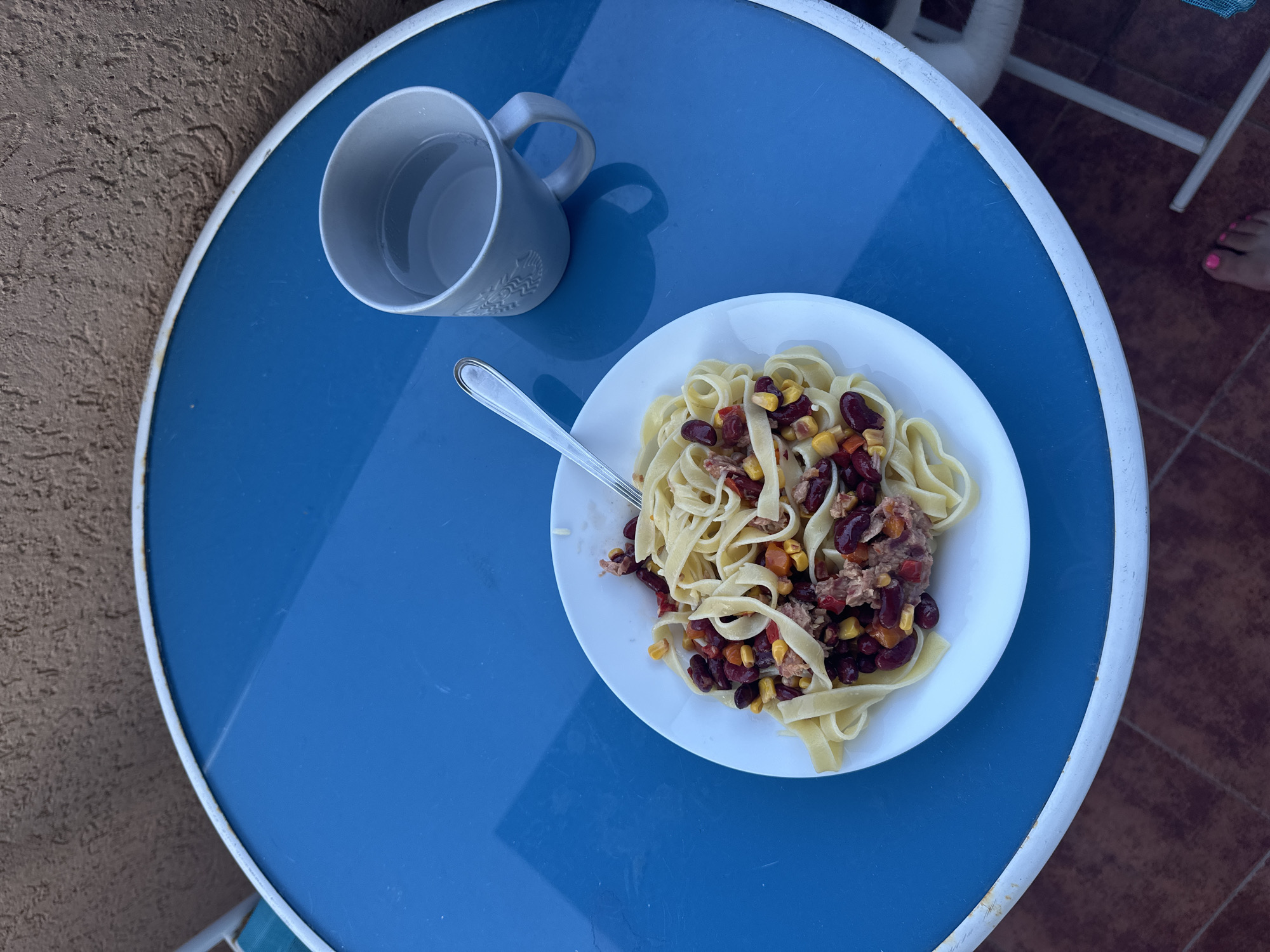 A plate of pasta with kidney beans, corn, and tuna is served on a blue circular table. The pasta appears to be fettuccine. Next to the plate is a grey mug filled with a clear liquid, likely water. The table is outdoors, positioned on a reddish-brown tiled surface. A textured beige wall is visible to the left of the table. The lighting suggests it is daytime, possibly late afternoon due to the shadows cast on the table. In the background, a white metal chair is partially visible, and a person's foot with pink toenails is near the edge of the frame. The setting appears to be a casual outdoor dining area, possibly a patio or balcony. There is no visible text in the image.