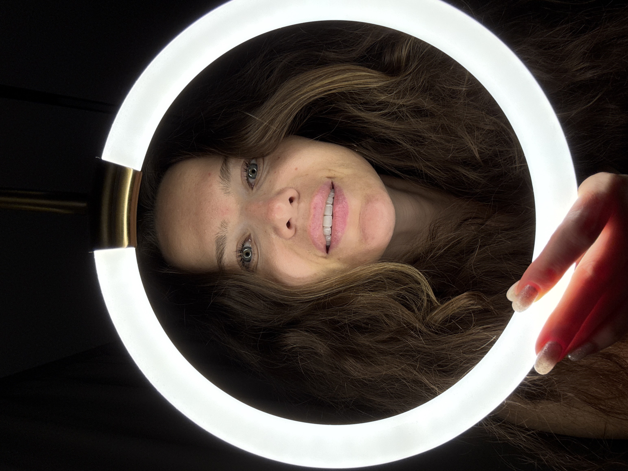 A close-up, high-angle, portrait-style shot focuses on a woman's face framed by a bright, circular light source, likely a ring light. The woman has long, wavy brown hair that falls around her face and shoulders. She has fair skin, light green eyes, and her lips are slightly parted, revealing her teeth. She is wearing subtle makeup, and her fingernails are visible in the foreground, illuminated by the light, appearing to have a glittery polish. The background is dark and indistinct. The ring light, a common tool for photography and videography, is the most prominent object in the image, creating a bright halo effect around the woman's face. The scene is set indoors, with the dark background suggesting a controlled lighting environment. There is no information in the image to suggest a location in Khimki, Russia.