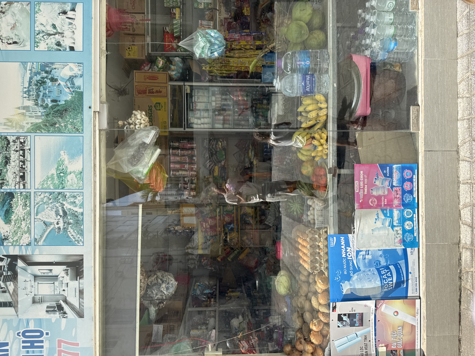 The image captures a street-level view through the glass window of a small convenience store, likely located in Bac Nha Trang Ward, Vietnam, as indicated by the presence of Vietnamese text on some products and advertisements. The reflection on the glass suggests an outdoor setting, possibly a street with buildings.
Inside the store, shelves are stocked with various goods. On the bottom shelf, there is a display of fresh produce, including potatoes, onions, a head of cabbage, and a rack of eggs. There are also bottled beverages, including Aquafina water and possibly other drinks. To the right, there are more bottled drinks on display.
Prominently featured at the front of the store, at eye level, are several large product advertisements. One is a bright blue box advertising "Pocari Sweat," a rehydration drink. Next to it is a pink advertisement for "Yakult," a probiotic drink. Other advertisements and product boxes are visible, some featuring images of people and product branding.
In the background, various shelves are filled with packaged food items, canned goods, and other miscellaneous products. The lighting inside the store appears to be a mix of artificial and natural light, creating reflections on the glass. A person is partially visible inside the store, appearing to be a customer or employee, holding a bouquet of flowers and looking at their reflection. Another person, appearing to be a woman, is standing further inside the store, facing away from the viewer. The overall scene depicts a typical, modestly stocked neighborhood store.