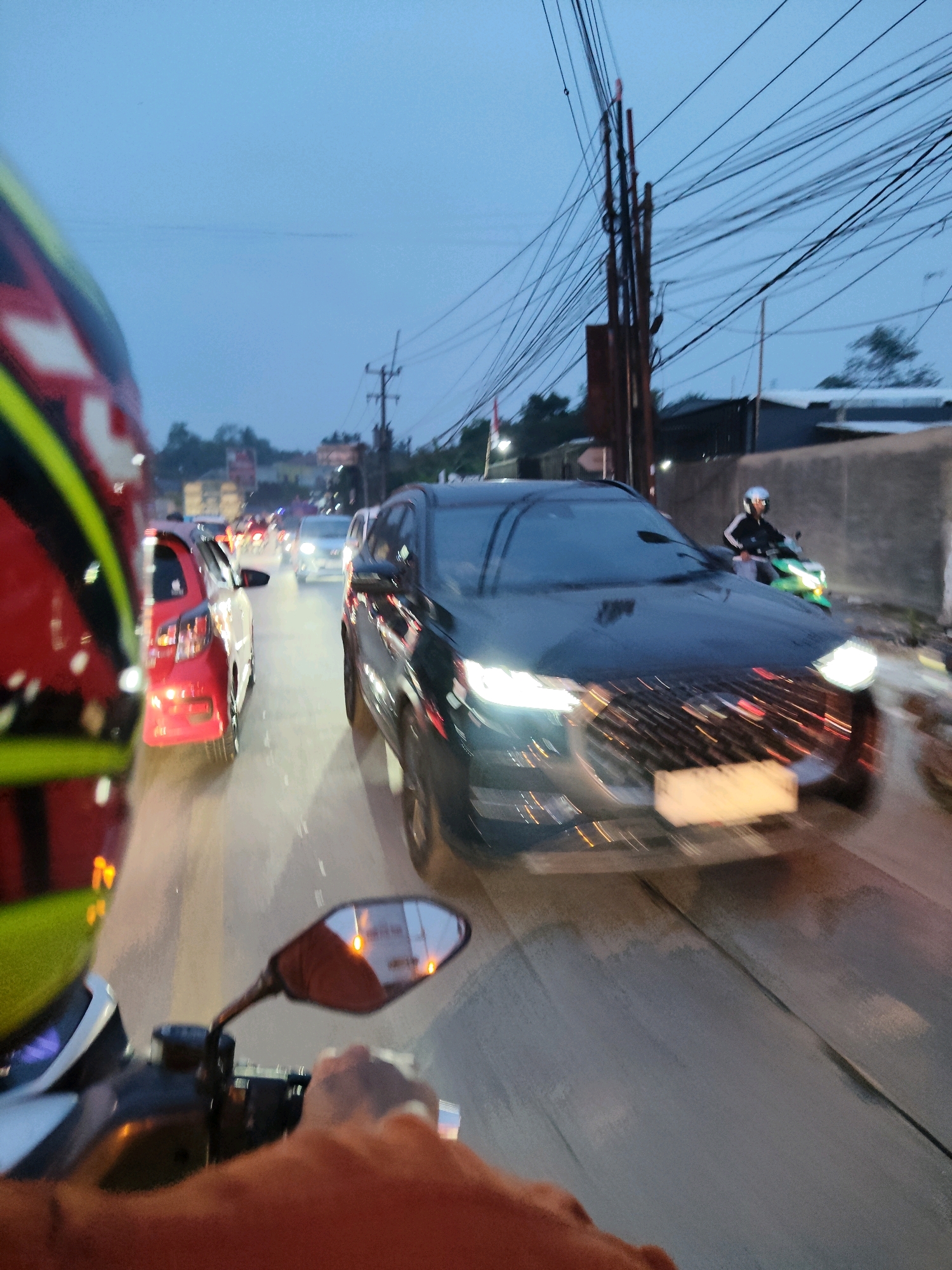 The image depicts a street scene in Kaduagung Tengah, Indonesia, at dusk or twilight. The perspective is from a motorcycle, with the rider's orange shirt and hand visible in the foreground, holding the handlebar. A motorcycle helmet, red and yellow, is partially visible on the left. The road is wet, suggesting recent rain, and traffic is heavy. Several cars and motorcycles are present. A black SUV is prominent in the center, with its headlights illuminated. A red car is to its left. Further down the road, other vehicles with illuminated headlights can be seen. A person on a green motorcycle is visible to the right of the black SUV. Overhead, numerous power lines crisscross the sky, supported by utility poles. Buildings and trees are visible in the background. The overall atmosphere is that of a congested urban street during low-light conditions.