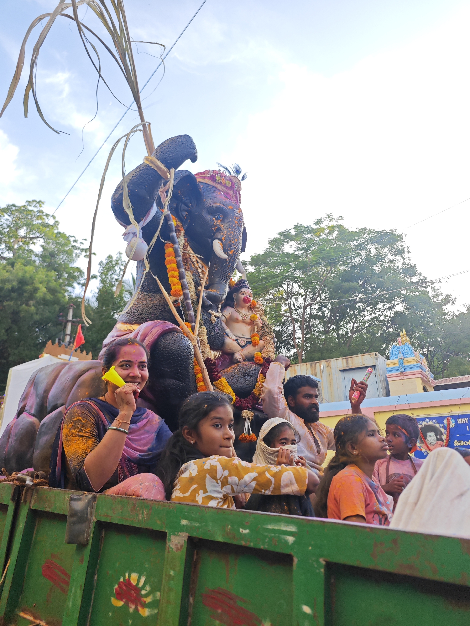 This is a medium shot, eye-level, outdoor photograph taken in Puttaparthi, India, likely during a festival, given the presence of colored powder on people's faces and clothing, and a large decorated elephant. The primary subject is a large, dark-colored elephant statue, adorned with garlands and a headdress, carrying a smaller statue of a deity on its back. Several people are seated in the back of a green, open-top truck, in front of the elephant.
In the foreground, a woman with dark hair and a blue and pink garment is smiling and holding a yellow object to her mouth, possibly a whistle or noisemaker. She has colored powder on her face and shoulders. To her right, a young girl with long, dark hair wears a yellow and white patterned shirt and looks to her right. Further to the right, a young girl with her face covered by a white cloth looks towards the elephant. Behind the woman, a man with a beard and dark clothing looks forward, holding a pink object. To his right, another young person in a pink shirt looks to their left, and a child in a pink shirt with a red bandana on their head is looking forward. A white cloth or garment is visible on the far right.
The elephant statue is very detailed, with visible tusks and decorated ears. The deity statue on its back appears to be small and adorned with jewelry and garlands. The truck bed is green with red and yellow painted designs. In the background, trees and a building with a colorful, multi-tiered roof, typical of Indian temple architecture, are visible. The sky above is bright blue with some clouds. The overall atmosphere suggests a vibrant and celebratory event.