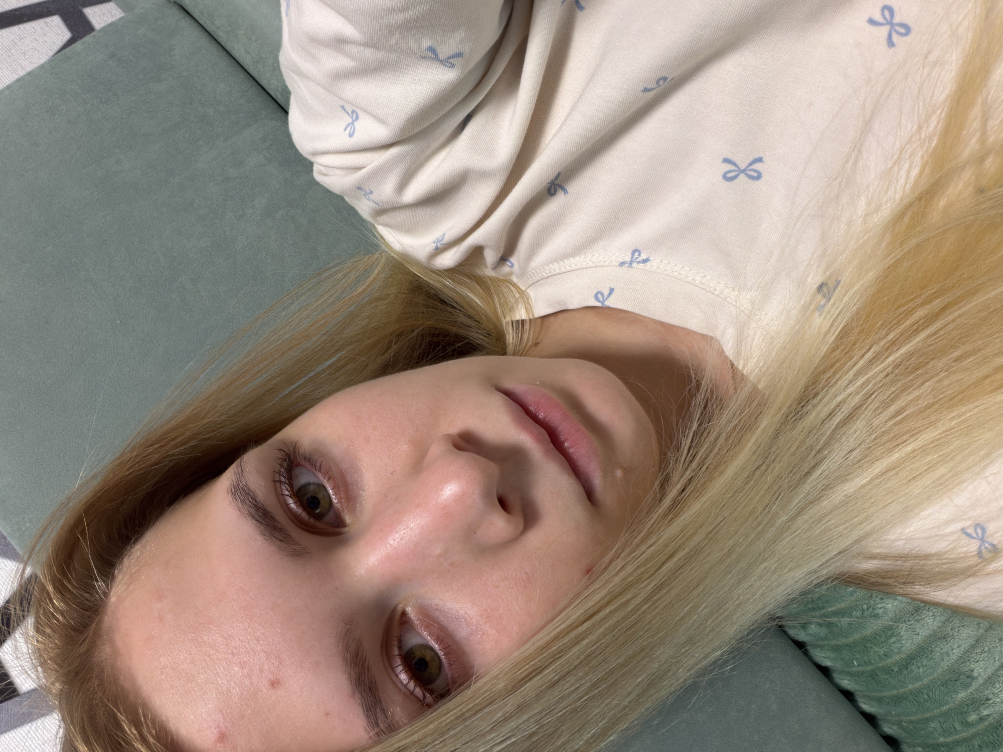A young woman with blonde hair is lying down and looking at the camera. She is wearing a light-colored t-shirt with small blue bows printed on it. Her hair is long and wavy, and it covers part of her face. The background is a blurred green surface, possibly a couch or a pillow. The lighting is soft and natural. The image appears to be a selfie. There is no discernible information about the location being Izhevsk, Russia.