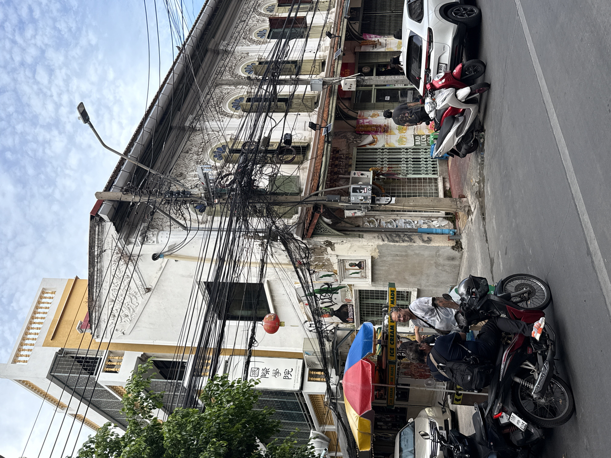 The image depicts a street scene in Bangkok, Thailand. In the foreground, several parked motorcycles line the side of the street, with two men standing nearby and interacting with each other. The man closest to the camera is wearing a white patterned shirt. The street is narrow, and the buildings along it appear somewhat old and ornate, with visible signs of weathering. A jumble of overhead power lines dominates the view above the buildings, typical in many parts of Bangkok. A red paper lantern hangs from one of the lines. Signage in Thai and English is visible, including "ICE CREAM HOME MADE" and "BAD LUCK." A white car is parked further up the street. The sky is partly cloudy, indicating daylight.