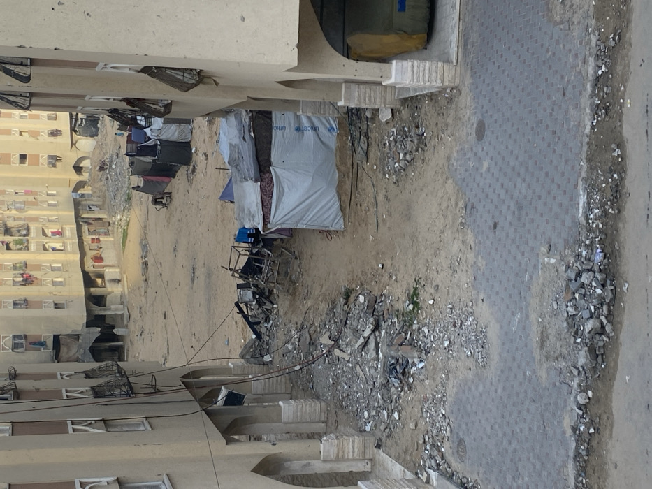 The image depicts an open urban landscape in Khan Yunis, Palestinian Territory, bordered by multi-story residential buildings. In the foreground, a paved sidewalk with grey interlocking pavers transitions into a rough dirt path heavily strewn with concrete rubble and assorted debris.
The central midground is an expansive, disturbed area of bare earth and sand, containing substantial piles of twisted metal frames, broken concrete, and other construction debris. To the right of this expanse, a cluster of makeshift shelters constructed from tarpaulins and fabric is visible. One prominent shelter on the right is fashioned from a light-colored tarp, clearly displaying the blue UNICEF logo. Further back, a row of additional, darker shelters extends. Two indistinct human figures are faintly visible in the deeper midground, one near the darker shelters and another further right near a cart-like object.
The background is dominated by multiple multi-story residential buildings with tan or light brown facades, framing the open area. These structures exhibit signs of wear, with some balconies holding items, including laundry, and windows fitted with metal grates. Ground floors feature arched openings. Dark electrical cables are seen descending from the upper levels of the building on the left. An arched opening on the right building reveals a large, yellow, tarp-covered object. The overall environment is dry and dusty, indicating disrupted ground conditions and makeshift living arrangements amidst permanent structures under natural daylight, evidenced by long shadows.