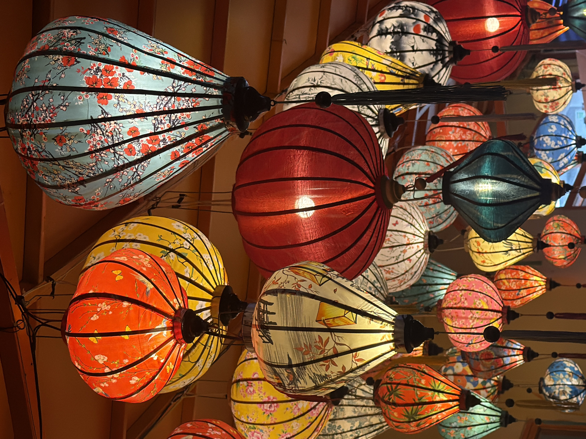 This media file captures a vibrant and densely packed display of numerous illuminated traditional Vietnamese lanterns, showcasing a rich tapestry of colors, patterns, and shapes. The lanterns vary significantly, with prominent hues including red, teal, yellow, orange, pink, white, and blue, each glowing from an internal light source.
Many lanterns feature intricate designs such as cherry blossoms, other floral patterns, birds, butterflies, and what appear to be subtle landscape elements or traditional motifs. Shapes range from rounded and spherical to elongated and tapered, often with visible black or dark-colored ribs forming their structure. Several lanterns are adorned with tassels or decorative strings hanging from their bases.
The lanterns are suspended at different heights, creating a layered and immersive canopy effect overhead, set against a warm, wooden-toned ceiling or wall background. The bright illumination suggests an evening or nighttime setting, or an indoor space where artificial light is dominant, creating a festive, warm, and inviting ambiance. The scene is consistent with cultural decorations found in Đà Nẵng, Vietnam.
No people or visible text are present in the image.
