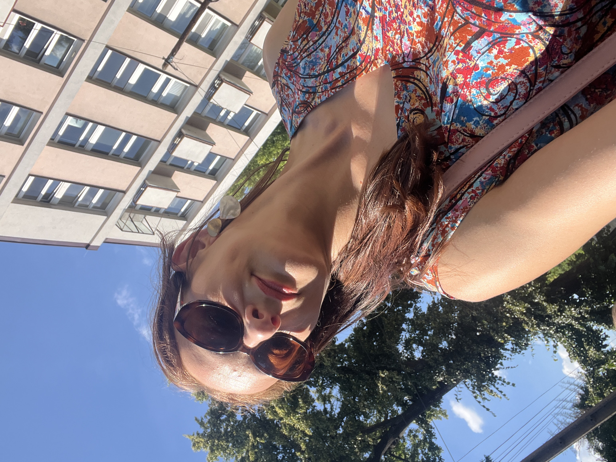 A woman, her face partially obscured by sunglasses, smiles at the camera in a close-up, low-angle selfie. She has long, dark brown hair and is wearing a colorful, patterned dress. A light pink strap, likely from a purse, is visible across her chest. The background features a modern building with multiple windows and a clear blue sky dotted with a few clouds. Lush green trees are also visible, suggesting an outdoor setting. The lighting indicates it is daytime, likely sunny, with the sun casting a shadow on the woman's dress. Her expression appears happy and relaxed. There is no visible text in the image.