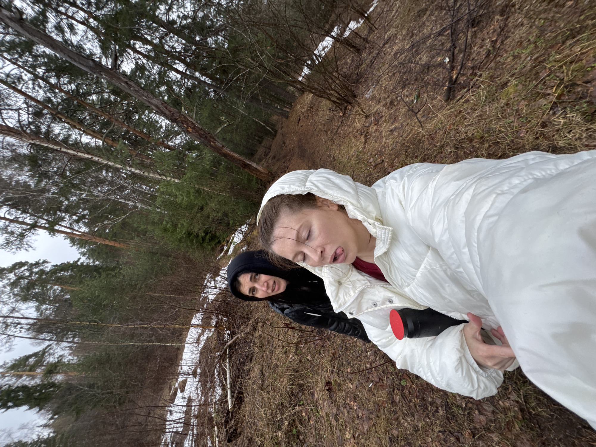 Two individuals are in a wooded area. One person is wearing a white, hooded puffy jacket and has their eyes closed and tongue slightly protruding. They appear to be holding a black cylindrical object with a red top. The second person is wearing a black hooded jacket and looking at the camera. The background consists of trees, dry foliage, and some patches of snow. The scene is outdoors, and the perspective is from a low angle. The location is Chaykovsky, Russia.