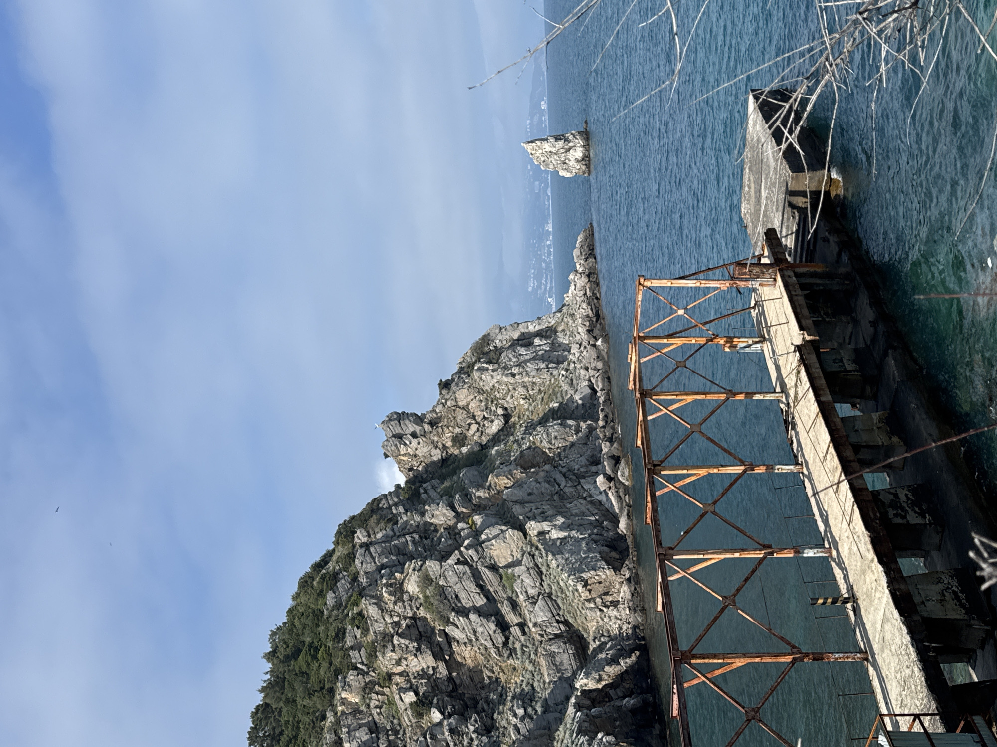 The image depicts a rocky cliff and a deteriorating concrete structure with rusted metal supports extending into the water, located near Ливадийский поселковый совет, Ukraine. The scene includes a view of the blue sea and sky, with a small rocky islet in the distance. The cliff is covered with vegetation at its base.