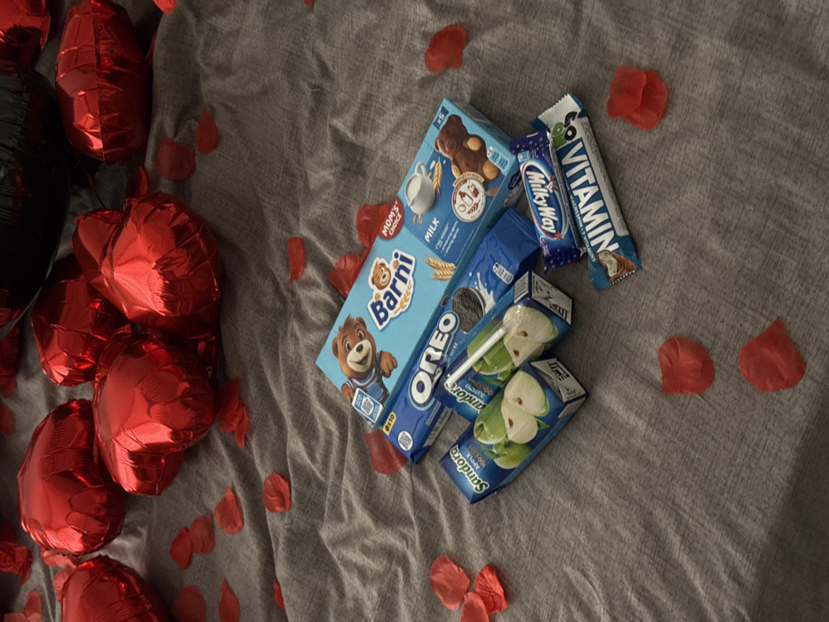 This image depicts an interior scene, likely a domestic setting within Tbilisi, Georgia. The primary surface is a textured, wrinkled grey fabric, resembling a bedspread or blanket. Scattered across this surface are numerous artificial red rose petals.
In the upper portion of the frame, several red, metallic, heart-shaped foil balloons are visible, some partially obscured. A portion of a black balloon is also visible at the top right.
A collection of packaged snack items is arranged centrally on the grey fabric. These items include:
* One light blue box of "Barny" mini-cakes, labeled "MOM'S CHOICE" and "MILK".
* One dark blue package of "Oreo" cookies.
* Two small carton boxes of "Sandora Apple 100% Juice," each depicting green apple slices. One juice box has a white plastic straw inserted into it.
* One "MilkyWay" chocolate bar, still in its wrapper.
* One "VITAMIN" chocolate bar, featuring the text "CHOCOLATE + VITAMINS" and an image of a coconut, also in its wrapper.
No individuals are present in the scene, and no explicit actions or interactions are depicted. The arrangement suggests a prepared display or gift.