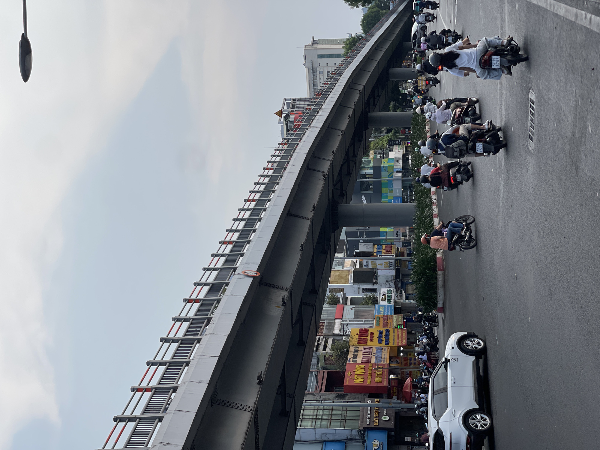The image depicts a busy street scene in Thủ Đức, Vietnam, dominated by an elevated concrete overpass. Below the overpass, a dense stream of motorcycles flows along the roadway. A white car is visible parked on the roadside. Signage in Vietnamese script is prominently displayed on storefronts lining the street. The sky is overcast. A streetlight stands tall on the left.