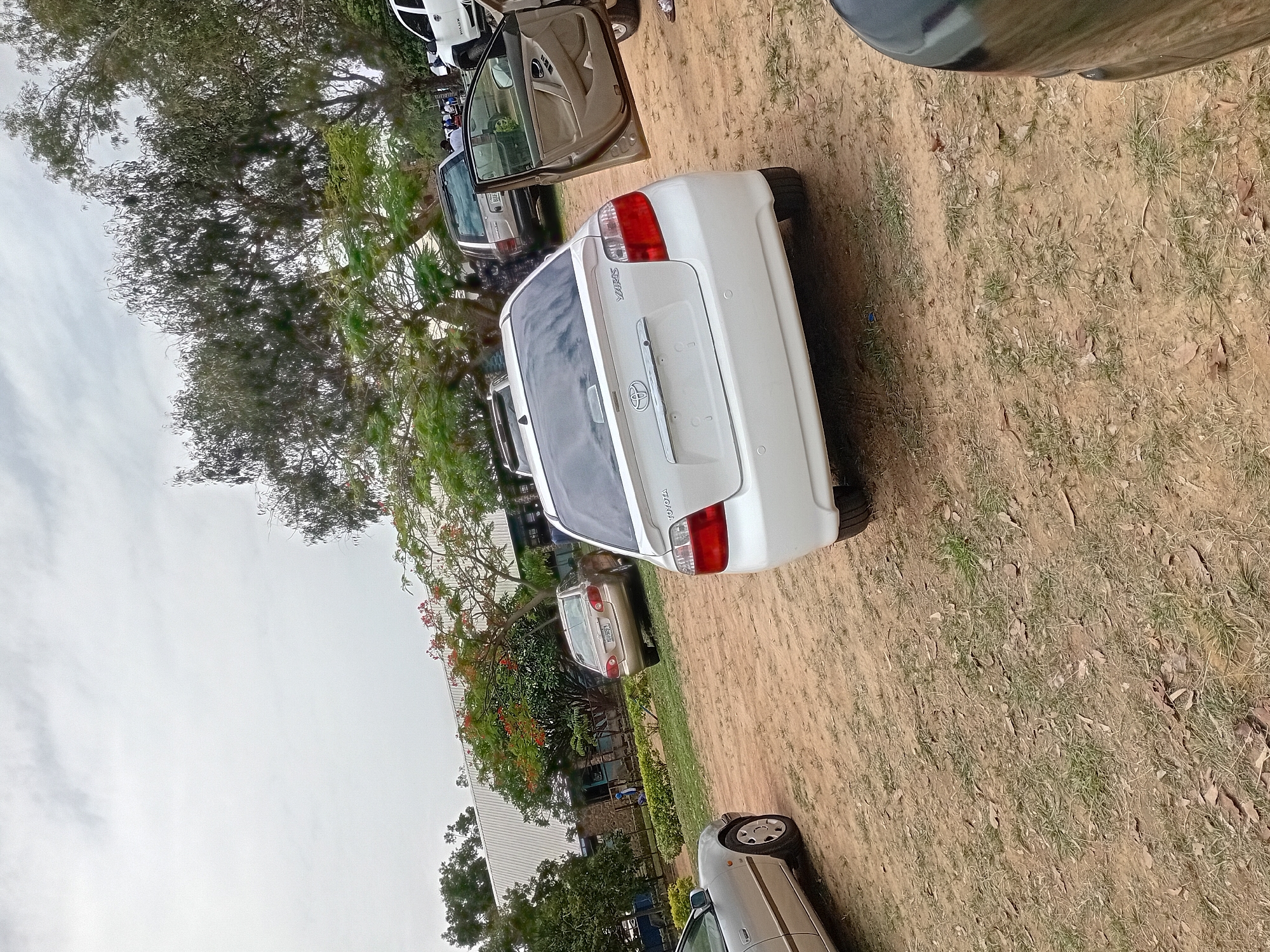 The scene depicts a collection of vehicles parked on a dirt lot. A white Toyota Yaris is prominently featured, its rear facing the camera. In the background, additional cars are visible, including a dark-colored vehicle with an open door, and other cars parked in rows. Trees and vegetation surround the parking area, some displaying red blossoms. The sky is overcast. This location is in the city of Jos, Nigeria.