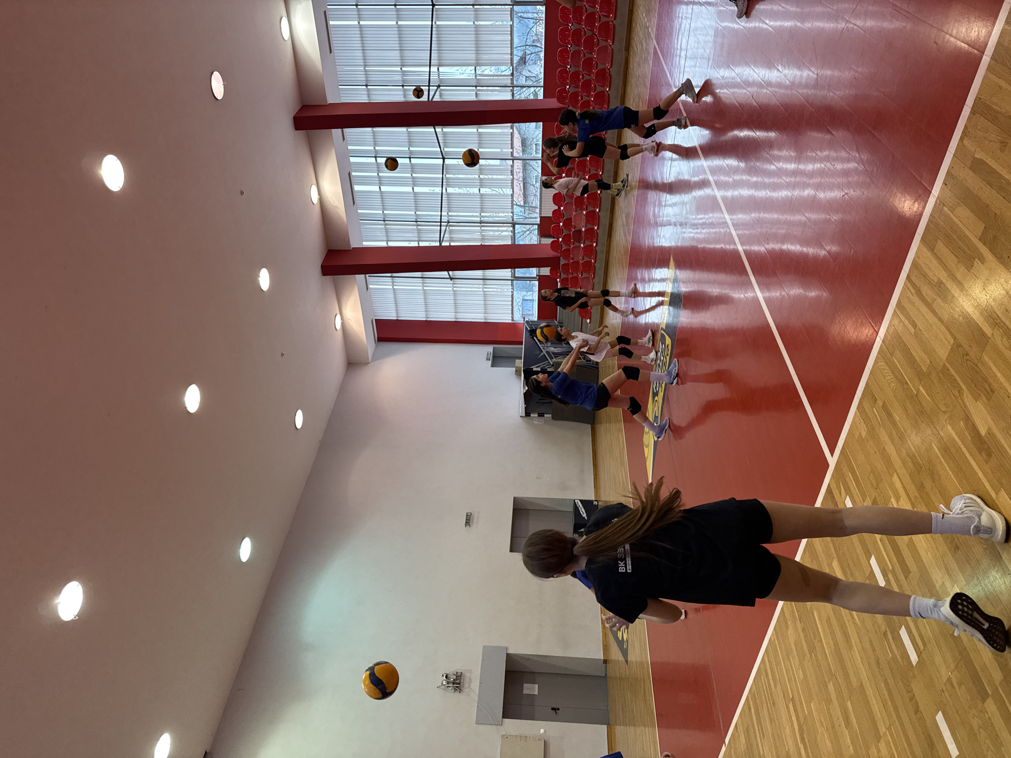 The image depicts a volleyball training session in a gymnasium in Etropole, Bulgaria. The gymnasium features a polished wooden floor with visible court markings, a white ceiling with recessed lighting, and red-painted sections on the walls and supporting columns. Seating is arranged along one side of the court.
Several young women are participating in a volleyball drill. One athlete is shown prominently in the foreground, preparing to hit a volleyball. Several other players are visible further down the court, spaced out in preparation for the drill. The players are wearing athletic gear including knee pads.