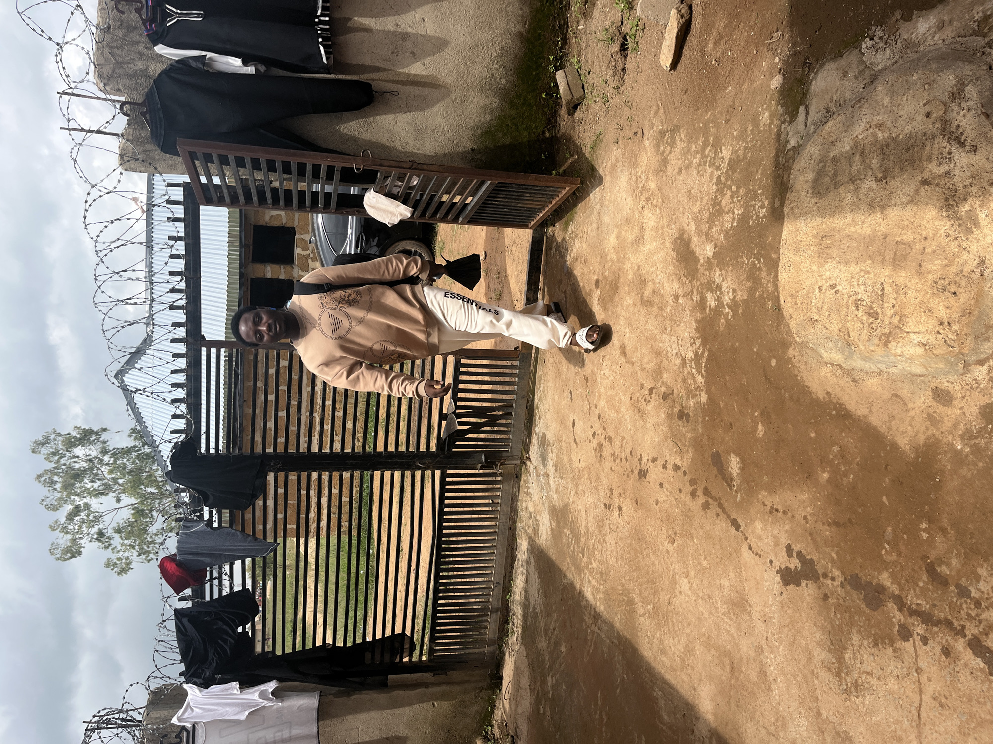 A young Black man stands outdoors in front of a black metal gate, possibly at the entrance to a residential compound. He is wearing a light brown hooded sweatshirt with a circular graphic design and white sweatpants with the word "ESSENTIALS" printed vertically in black letters. He is also wearing white slides. The man is looking towards the camera with a neutral expression.
The background features a wall with barbed wire on top and several pieces of clothing, including black t-shirts and a white shirt, hanging on what appears to be a clothesline or fence. Beyond the gate, a car is partially visible. The ground is covered in dirt and concrete. The overall scene suggests a casual, everyday setting. The lighting suggests it is daytime. The weather appears overcast. The provided location context places this scene in Durumi, Nigeria.