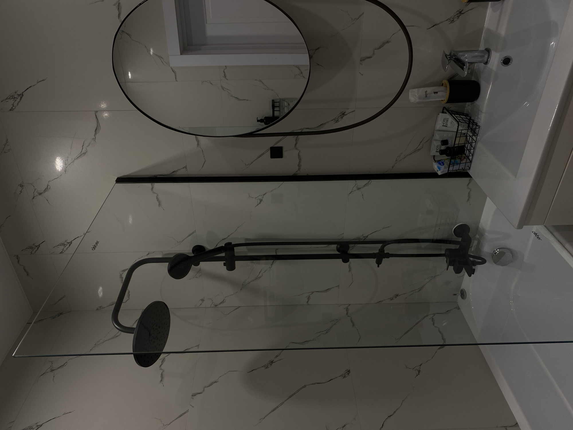 This image captures a modern and clean bathroom interior, likely within a hotel, as suggested by the amenities. The walls are adorned with large, glossy white tiles featuring a subtle grey marble-like vein pattern.
On the left side of the room is the shower area, enclosed by a clear glass screen. Inside, a contemporary black-finished shower system is installed, comprising an overhead rain shower head and a handheld shower connected by a black hose and fixtures. Faintly visible on the glass screen is the word "obbber."
To the right, a white ceramic sink with a chrome faucet is present. A black-framed circular mirror hangs above the sink, reflecting a window or door frame. Various toiletries are arranged around the sink: a black pump dispenser, white shampoo/conditioner bottles, and a black wire basket filled with individually wrapped "HOTEL SERIES" amenities like small bottles and soaps. Another black holder contains more "HOTEL" branded items. The brand name "obbber" is also faintly visible on the rim of the sink.
The room is well-lit, highlighting the clean surfaces and minimalist design. No people are visible, and no specific activity is taking place; the scene is static, showcasing the bathroom's setup. The overall aesthetic is one of contemporary elegance.