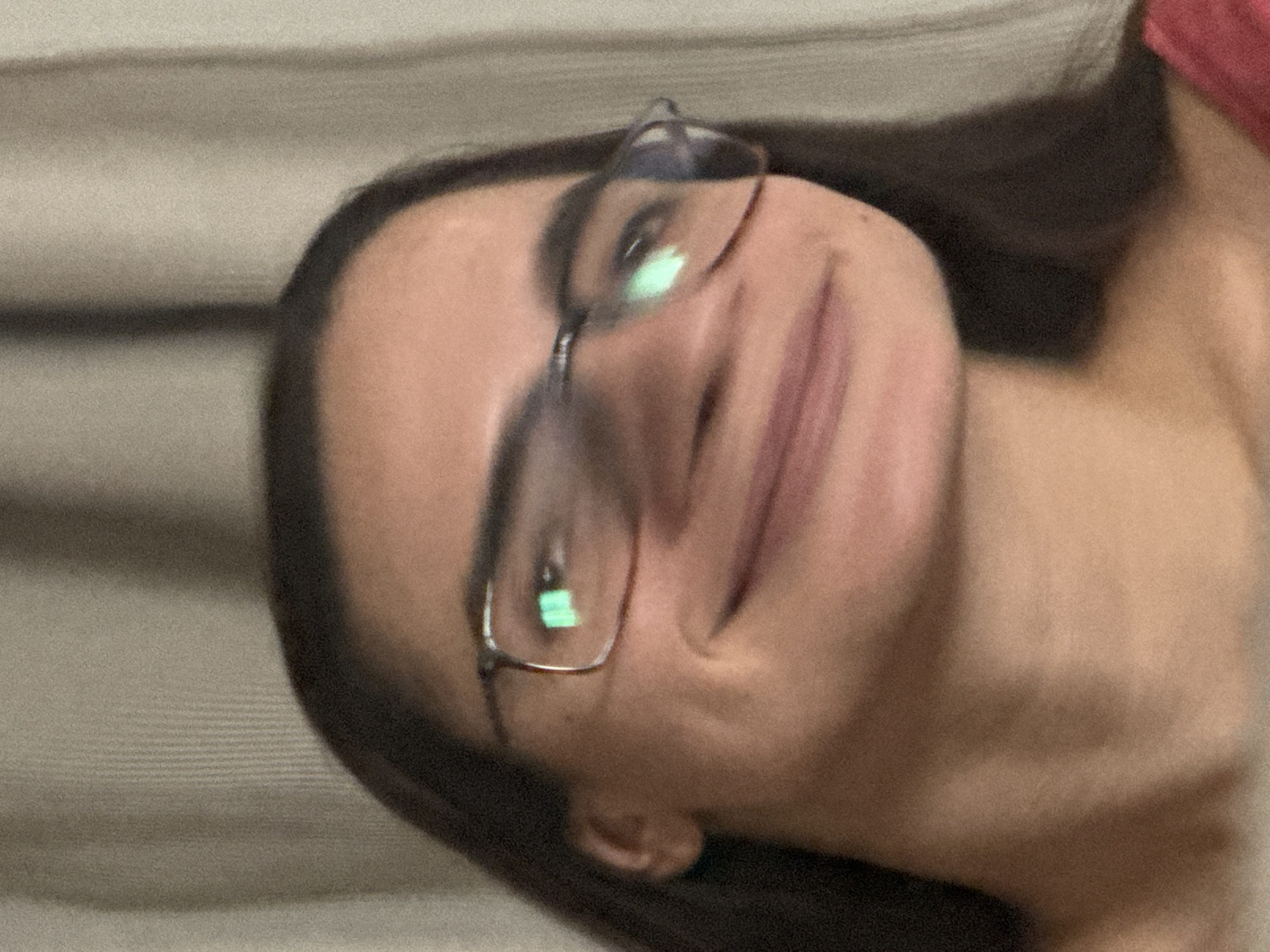 A close-up, slightly tilted image shows a woman with brown hair and glasses, smiling gently. She is positioned in front of what appears to be a textured, light-colored background. The scene doesn't provide any information or cues to determine its location in Krasnoyarsk, Russia, or any specific details about her actions or interactions with the environment.