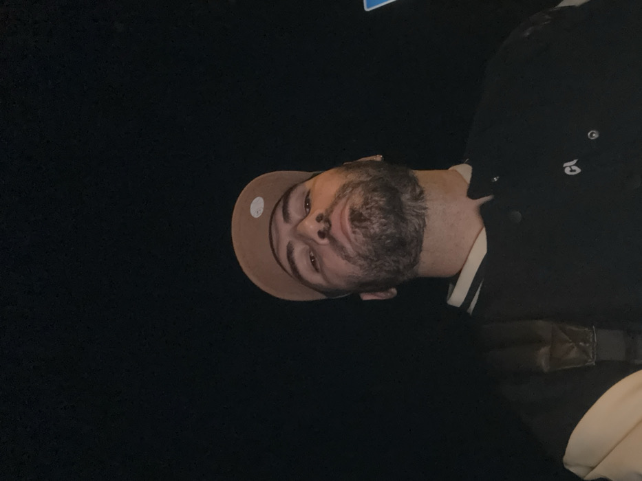 A man with a light brown baseball cap faces the camera against a dark background. His beard is graying, and he wears a dark jacket with white trim. The image is rotated, placing the man in the upper-right quadrant of the frame. The lighting is dim, obscuring details of the man's clothing and the surrounding environment.
