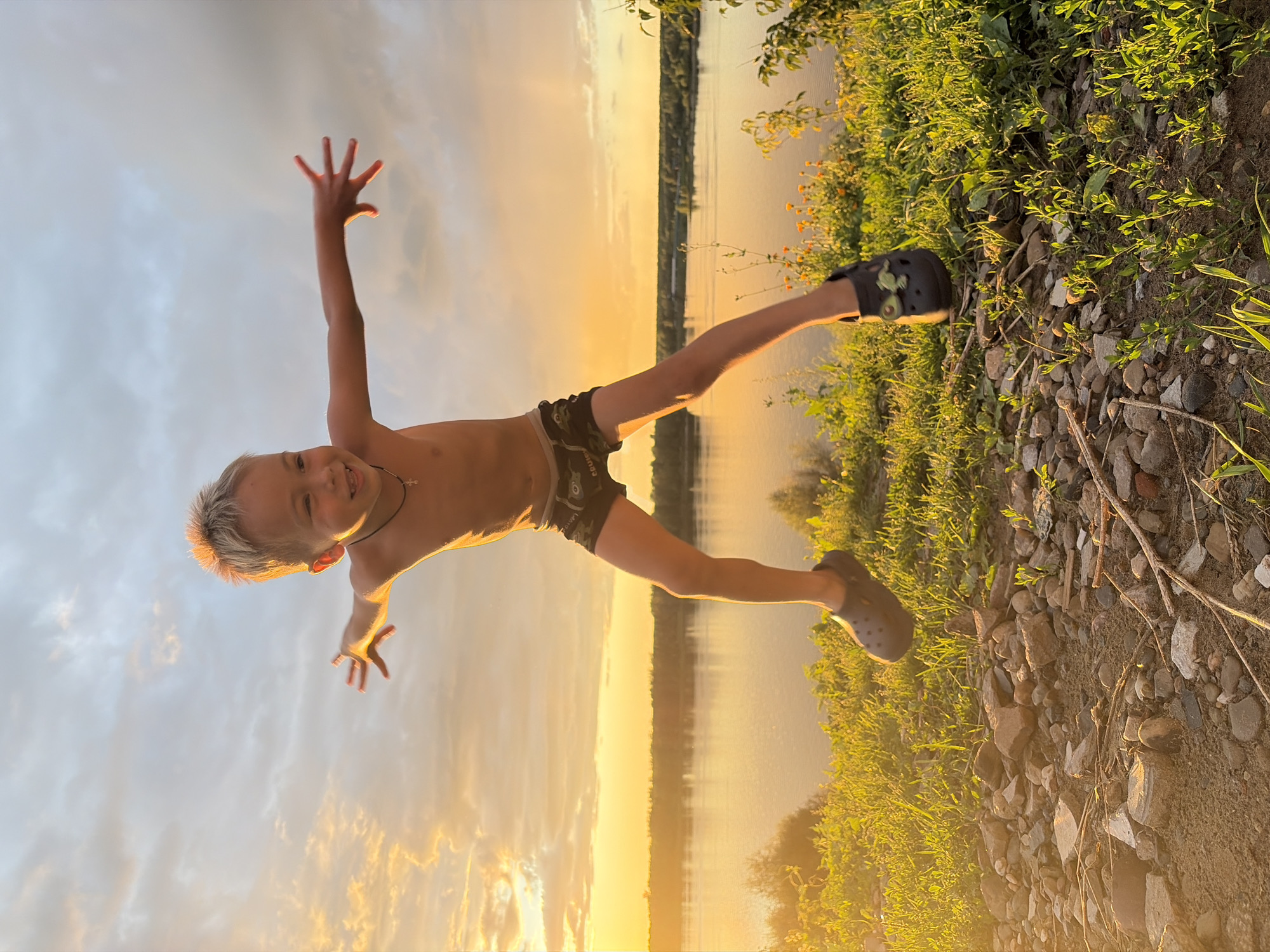A young boy with blond hair is captured mid-air, jumping with his arms outstretched to the sides. He is wearing dark patterned shorts and dark grey crocs. He has a faint necklace with a cross pendant. The boy appears to be smiling and looking towards the camera. The scene takes place outdoors during sunset, with a warm, golden light illuminating the sky and casting a glow on the boy and the surrounding environment.
In the background, a wide river stretches across the frame, reflecting the colors of the sunset. The riverbanks are lined with green trees and vegetation. The immediate foreground is a rocky and grassy embankment, with some dry twigs and small plants scattered across the ground. The sky is partly cloudy, with streaks of orange and yellow from the setting sun.
This scene is set in Olkhovka, Russia, a location known for its natural beauty, particularly its river and surrounding landscapes. The time of day is late afternoon or early evening, as indicated by the sunset.