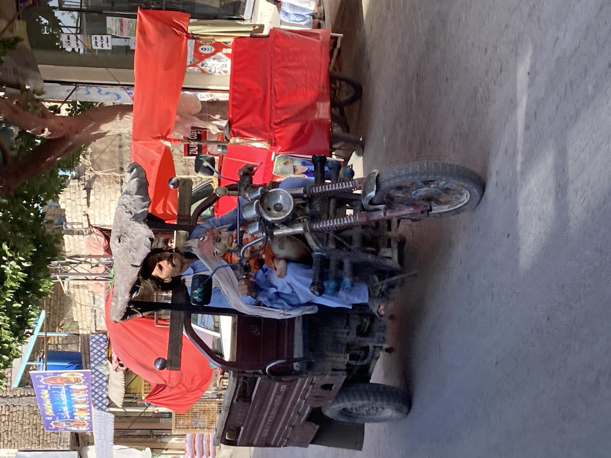 A man is seated in the driver's seat of a three-wheeled vehicle in Quetta City Tehsil, Pakistan. The vehicle has a red canopy and a weathered frame. The man is wearing a blue shirt and appears to be operating the vehicle. To the left of the vehicle, a food stall or shop is visible, with a menu board displaying images of food items in Urdu. The background shows a street with buildings. The image is taken at an angle.