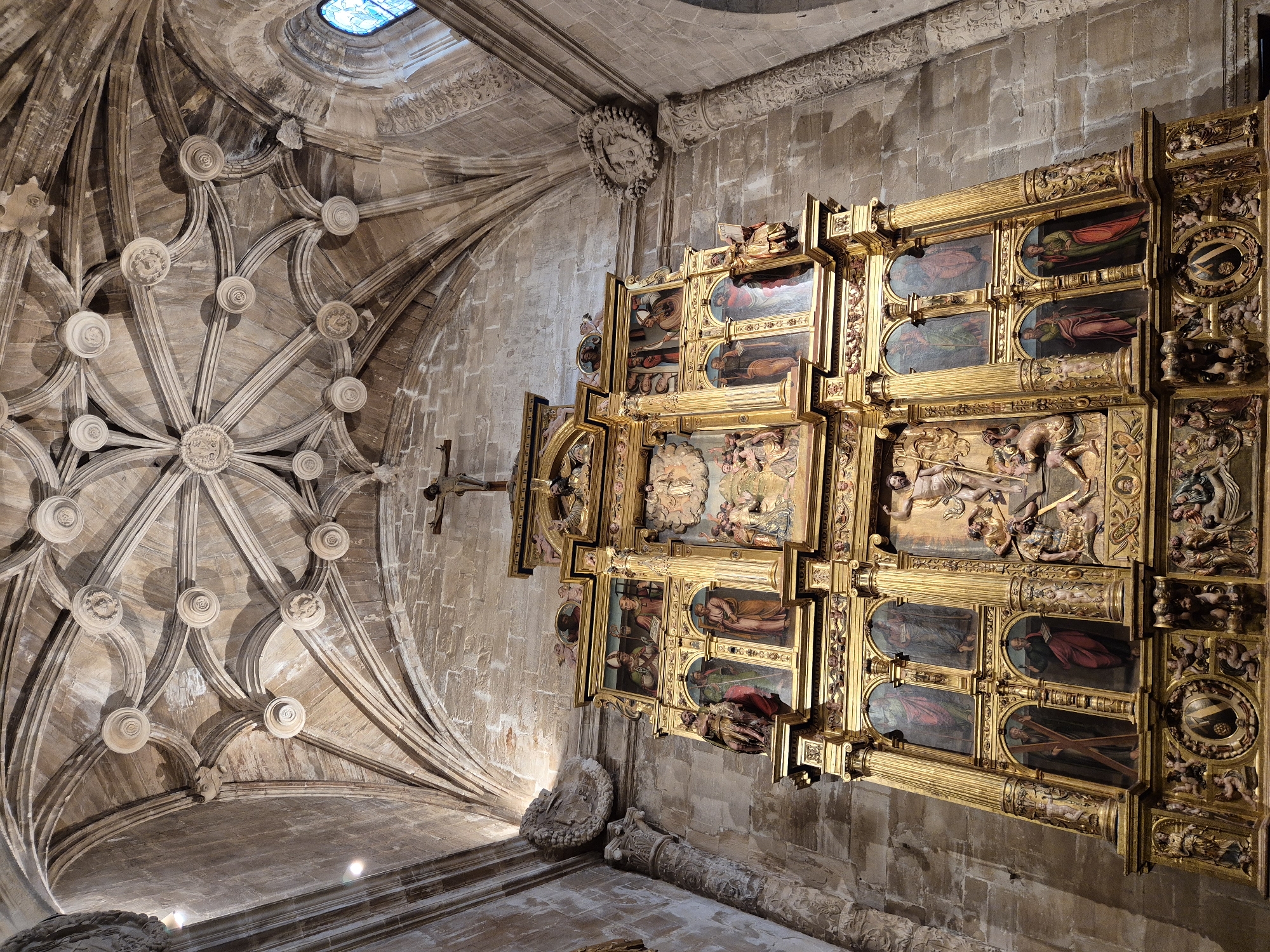 The image shows a gilded altarpiece within a stone church. The altarpiece is elaborately decorated with paintings and sculptures depicting religious scenes and figures. A crucifix stands at the very top of the structure. The ceiling of the church is vaulted, with intricate ribbing and decorative bosses. The stone walls are visible throughout the space.