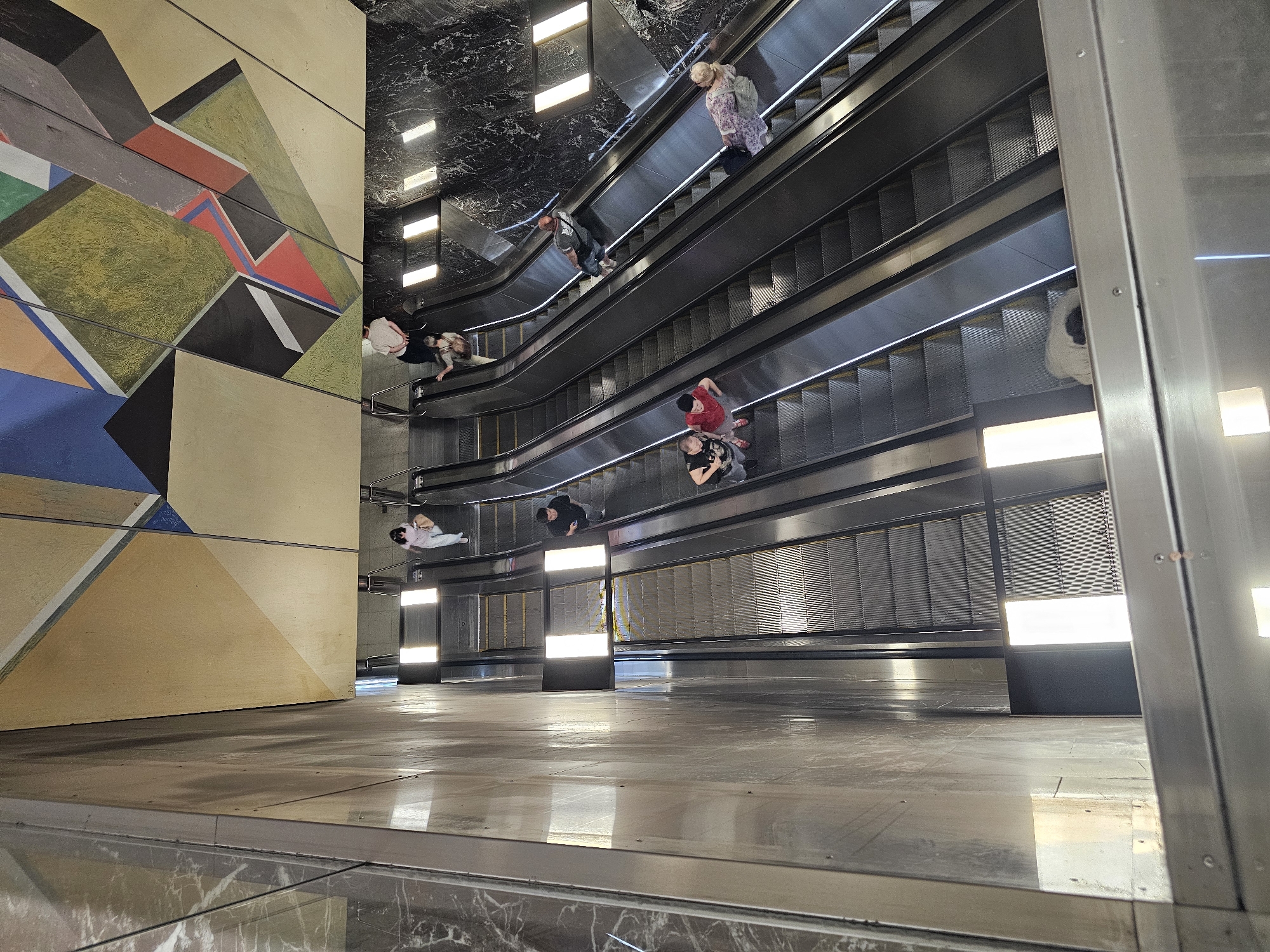 In a wide-angle, overhead shot of a modern escalator bank, several individuals are seen ascending and descending. The escalators are made of stainless steel, with illuminated steps and handrails. The background features a striking, abstract mural composed of geometric shapes in various colors, including yellow, blue, red, green, and black. The walls are clad in polished dark marble, and the overall environment suggests a public building, possibly a metro station or a commercial complex. The image is a high-angle shot looking down into the escalator well.