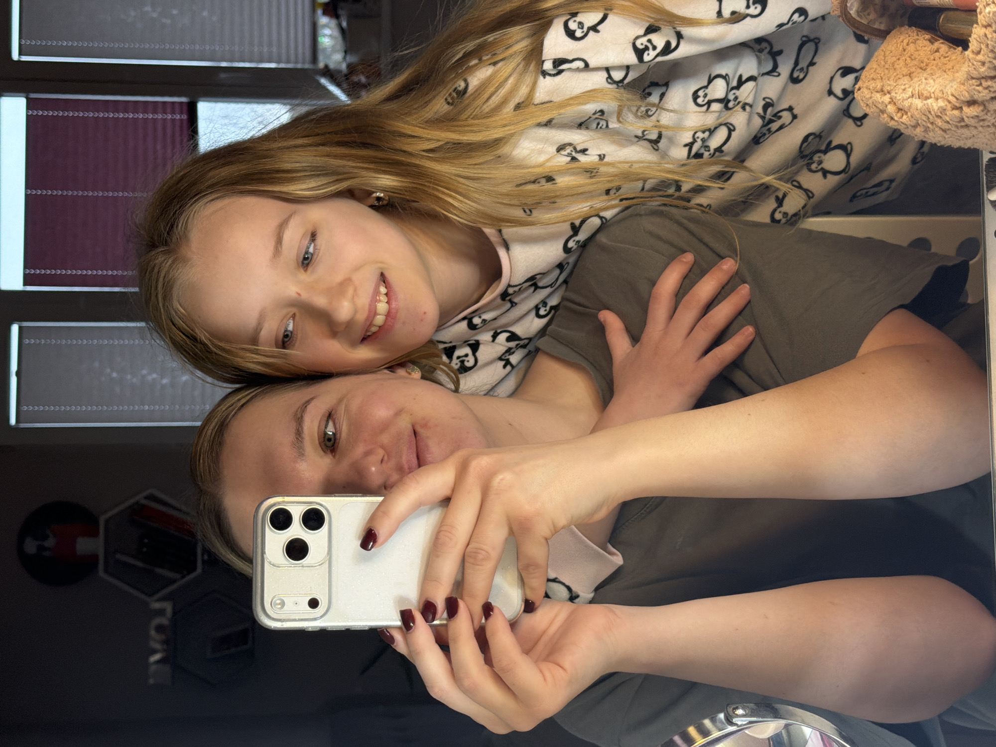 Two females are taking a selfie indoors. One female is older and holding a white smartphone with three rear cameras to take the photo. She has dark red nail polish on her fingers. The second female is younger and wearing pajamas with a panda bear pattern. They are positioned close together, with the younger female leaning on the older female. The reflection of a logo is visible on the glass surface in the background. Venetian blinds are visible through a window in the upper left of the frame. It is not possible to determine the exact location in Dreiliņi, Latvia, from this image.