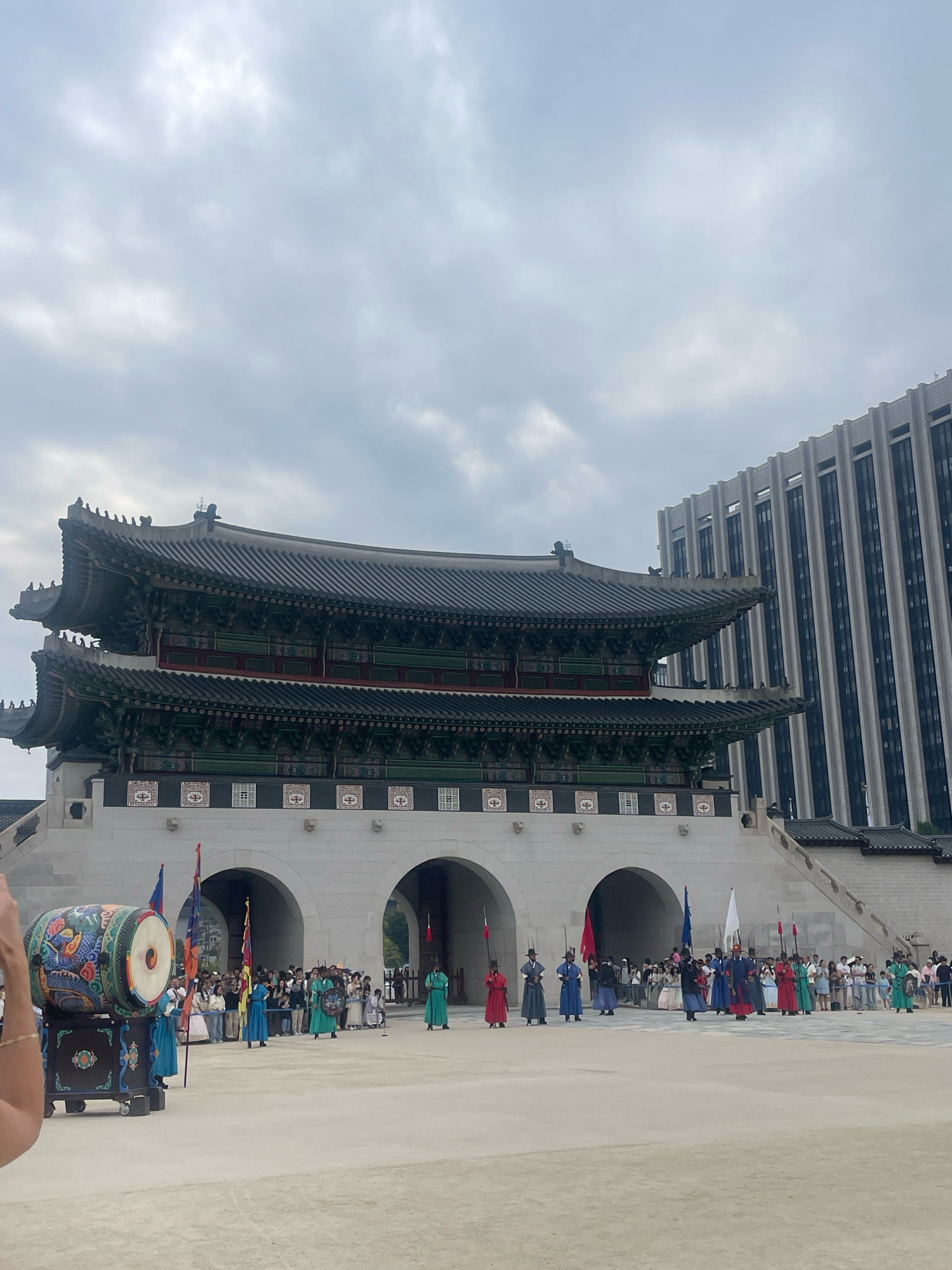 The image shows a scene at Gyeongbokgung Palace in Seoul, South Korea. In the foreground, a large, elaborately decorated drum is on display. Behind it, a group of people in traditional Korean hanbok attire are standing, some holding flags. Further back, the main subject is the historic palace gate, characterized by its tiered, dark tiled roofs and ornate architectural details. The gate is flanked by arched entrances and a stone staircase leading up. To the right of the palace, a modern, multi-story building with vertical lines is visible, contrasting with the traditional architecture. The sky is overcast with clouds. The scene appears to capture a cultural performance or historical reenactment.