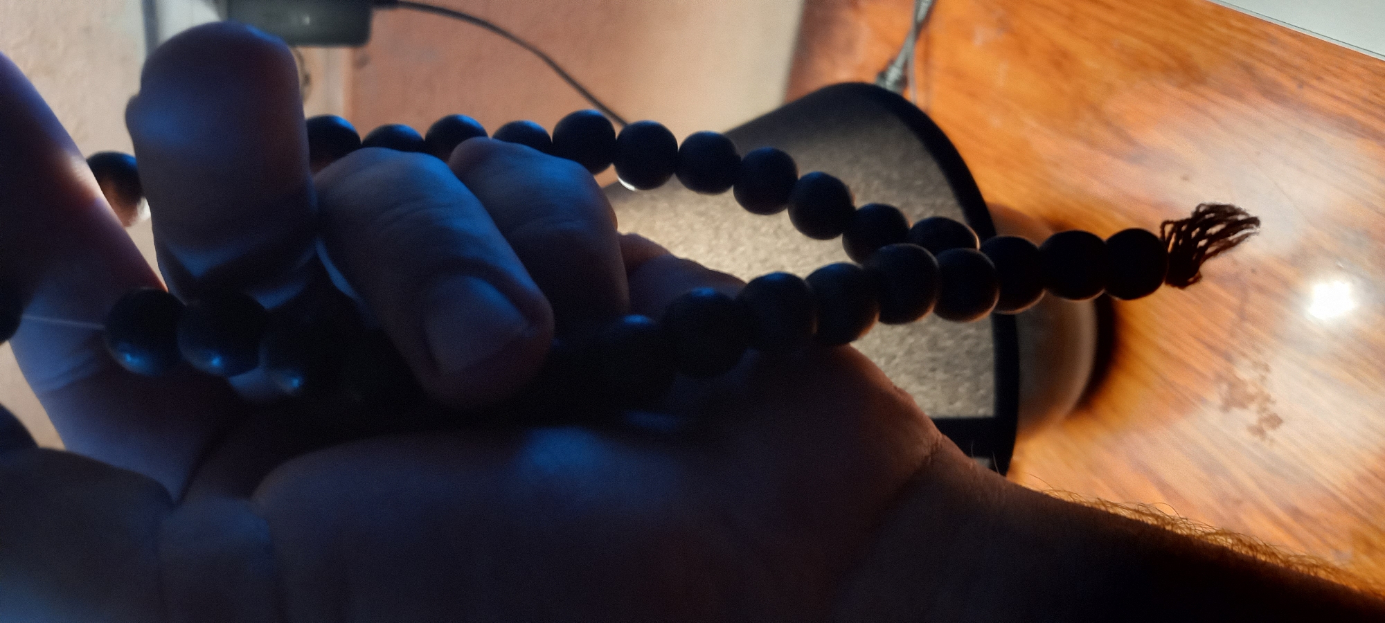 The image displays a close-up of a hand holding a string of dark, round beads. The hand and beads are illuminated by a blueish light, creating strong shadows. In the background, a wooden surface is visible, possibly a table, with a blurred object that resembles a lamp or a bowl. The lighting suggests an indoor setting with artificial light. There is no indication of the specific location within Kryvyi Rih, Ukraine.