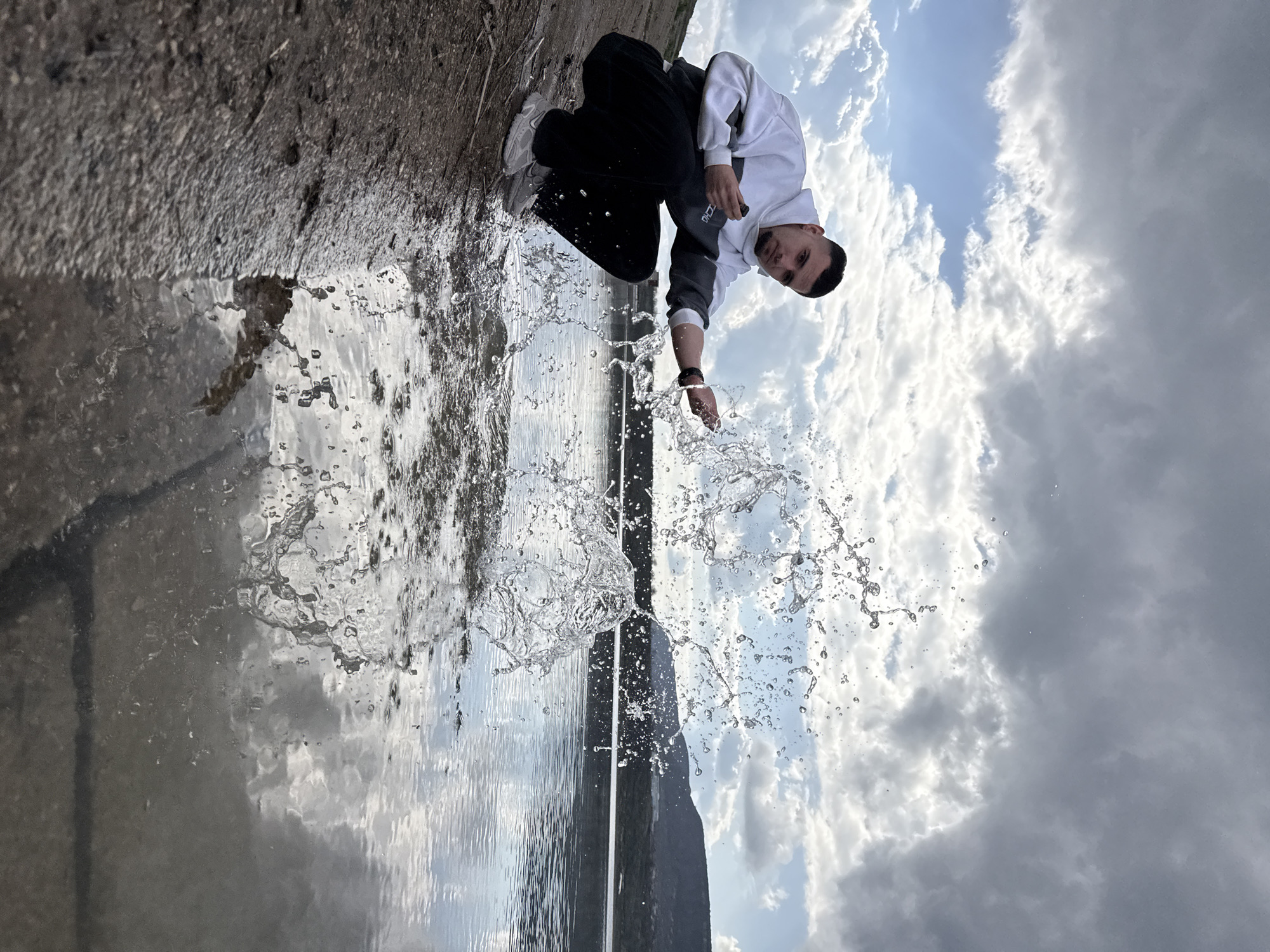 A man in a white and gray hooded sweatshirt and black pants crouches near a body of water. He is splashing water with his right hand, causing a spray of droplets. The background includes a cloudy sky and a concrete bank. The scene is likely located near a lake or reservoir in the city of Shumen, Bulgaria.