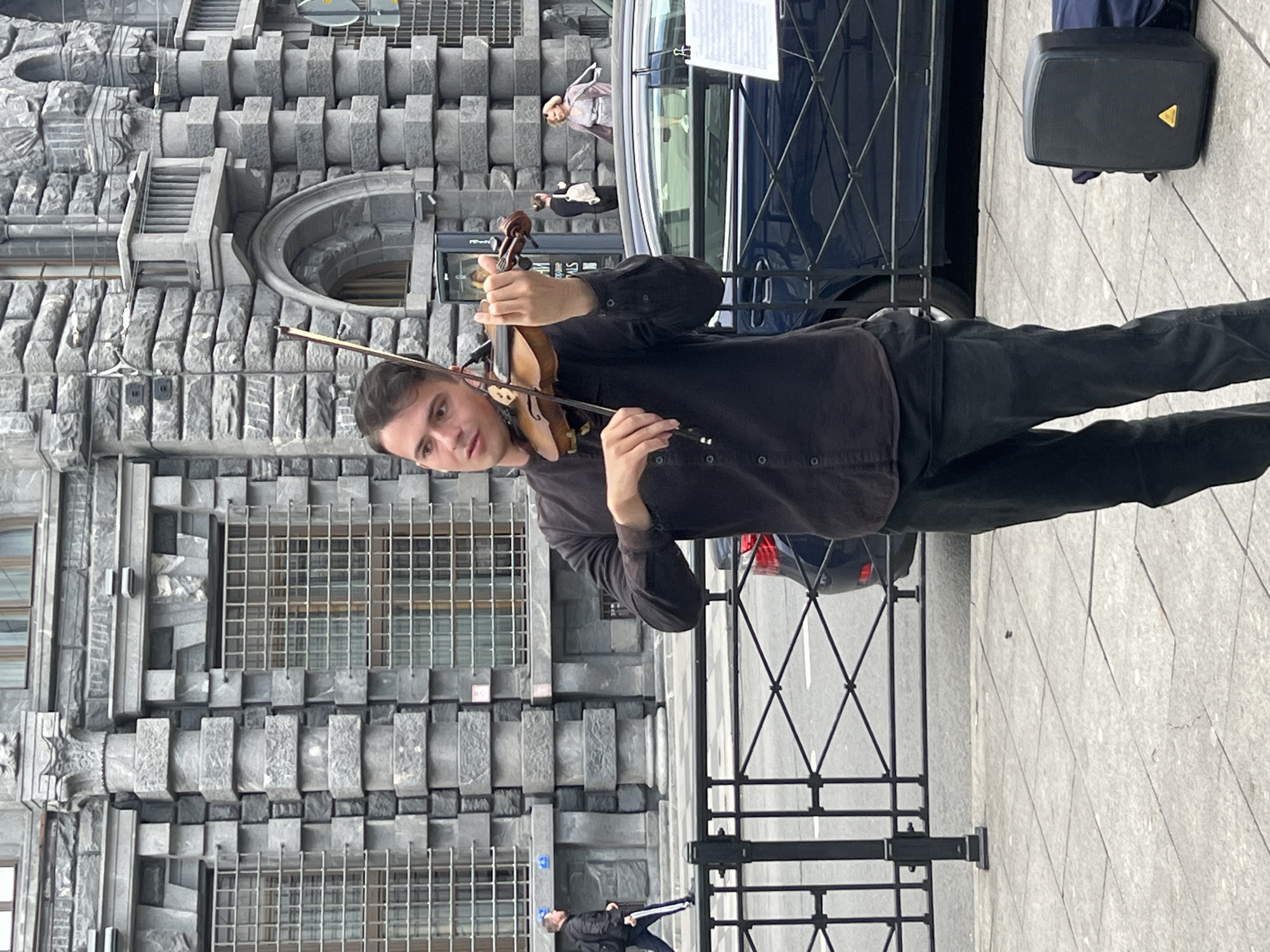 The image shows a man playing the violin on the sidewalk of a city street. He is dressed in dark clothing. Behind him, there is a distinctive building with a grey stone facade and a series of square blocks forming a patterned design. A car is partially visible behind him, and a black speaker bag is positioned on the pavement. People are walking on the sidewalk. The scene is likely located in Saint Petersburg, Russia, due to the architectural style of the building, which resembles the Eliseev Emporium on Nevsky Prospect.