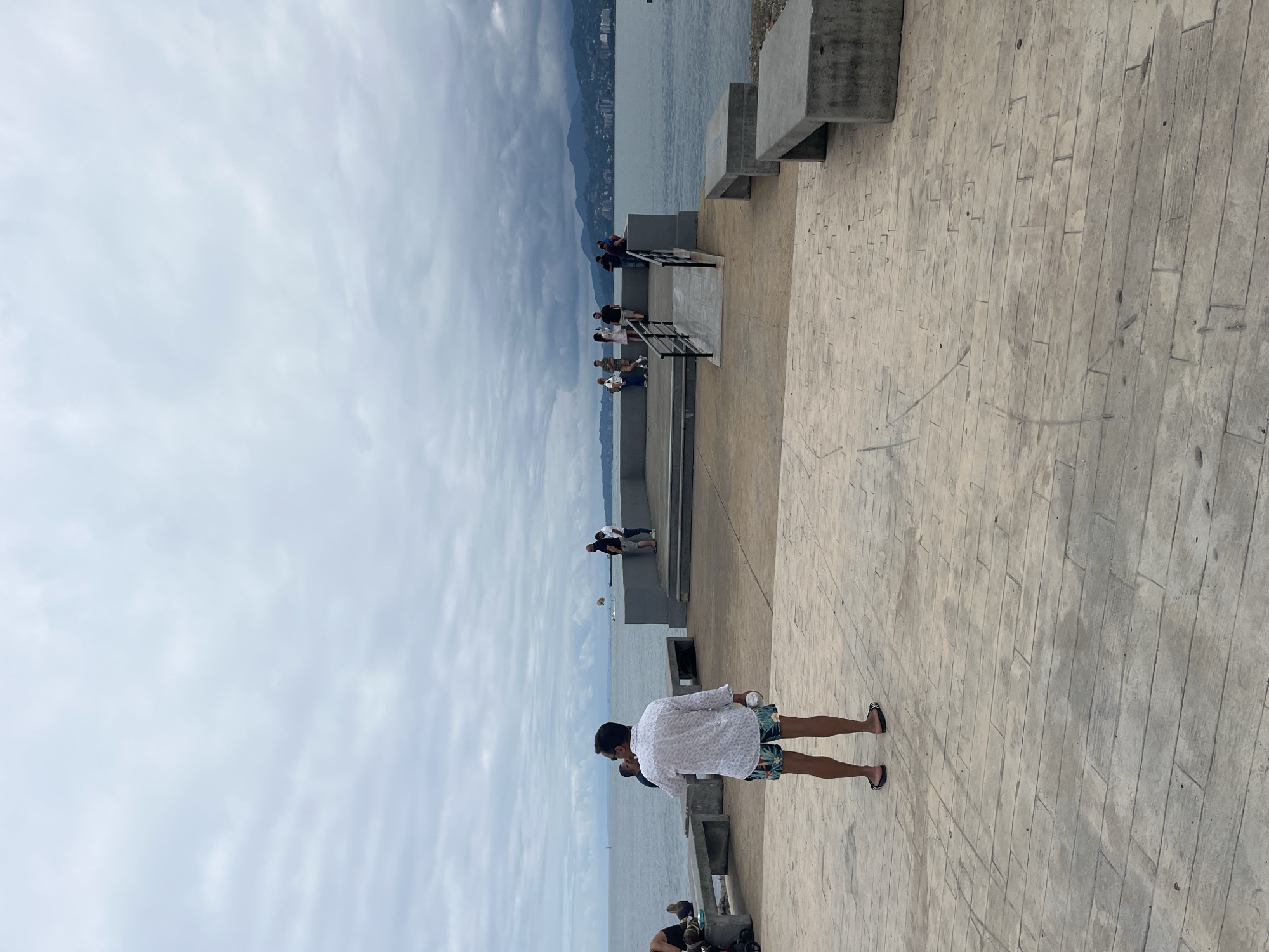 This is a full shot taken from a slightly elevated perspective of a paved promenade on a cloudy day. In the foreground, a man stands with his back to the camera, wearing a white patterned shirt and patterned shorts, holding a white bottle in his right hand. The promenade has a light beige, wood-like patterned surface. To the left and right of the promenade are long, grey concrete benches.
In the mid-ground, a raised concrete platform is visible, with a ramp leading up to it. Several people are gathered on this platform, appearing to be tourists. They are looking out at the sea, with some taking pictures. The sea is a calm, greyish-blue, and in the distance, a coastline with buildings and mountains can be seen. The sky is overcast with thick, grey clouds.
The location is identified as being in the city of Batumi, Georgia. The overall atmosphere is one of leisure and sightseeing, despite the dull weather.