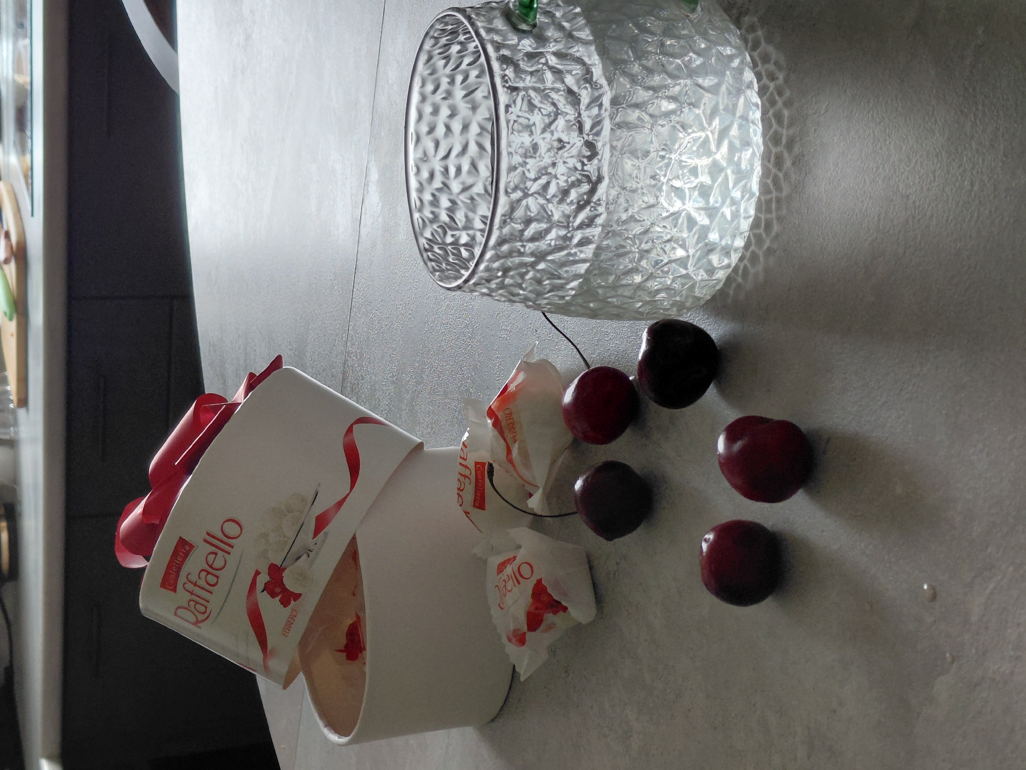 A still life photograph captures a set of RaffaELLO chocolates and cherries on a light gray countertop in a kitchen. A white cylindrical container of RaffaELLO chocolates, partially open and adorned with a red ribbon, is positioned on the left side of the frame. Several individually wrapped RaffaELLO chocolates, some appearing partially unwrapped, are scattered around the container. Five dark red cherries are also present, with one cherry stem visible near the container and another stem partially obscured by a wrapped candy. To the right of the chocolates and cherries is a clear glass tumbler with a textured, diamond-like pattern, filled with a clear liquid. The background shows a modern kitchen with dark cabinetry and a light-colored countertop. The lighting is somewhat dim, casting shadows on the countertop. The image appears to be taken from a slightly elevated perspective. There is no indication of the city of Novosibirsk, Russia, in the image.