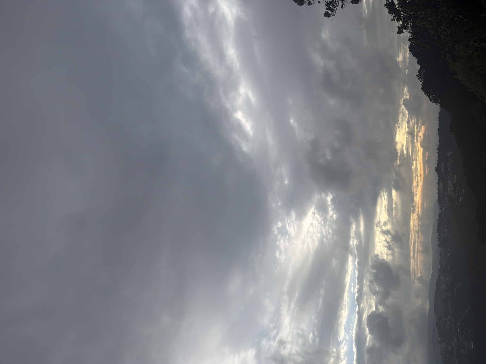 The image depicts a panoramic view of a cityscape nestled within rolling hills, likely at dawn or dusk, given the diffused sunlight breaking through a cloudy sky. The lower portion of the image is dominated by a densely populated area, with buildings of varying sizes and shapes creating a textured urban landscape. The hills surrounding the city are covered in dark, silhouetted foliage, some appearing as dense forests and others as scattered trees. The sky is filled with a dramatic display of clouds, ranging from heavy, dark grey masses to lighter, more wispy formations. Streaks of golden and yellow light emanate from the horizon, piercing through breaks in the clouds, suggesting the presence of the sun just below the visible horizon. The overall atmosphere is one of serene, natural beauty blended with human habitation, with the weather contributing to a moody and atmospheric scene. The location is identified as Xuân Thành, Vietnam.