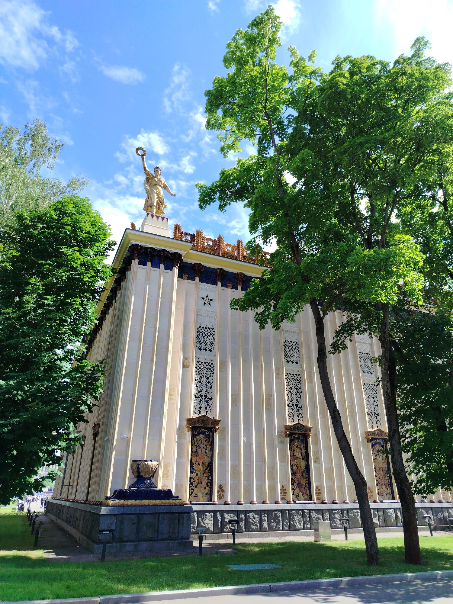 The image shows the iconic Pavilion of the Republic of Armenia at VDNKh in Moscow, Russia. The pavilion, a grand structure with a golden statue of a woman holding a wreath atop its roof, is adorned with classical columns and intricate decorative panels. Lush green trees surround the building, framing its elegant facade. The sky above is a vibrant blue, dotted with fluffy white clouds, suggesting a pleasant day. The scene conveys a sense of historical grandeur and architectural beauty.