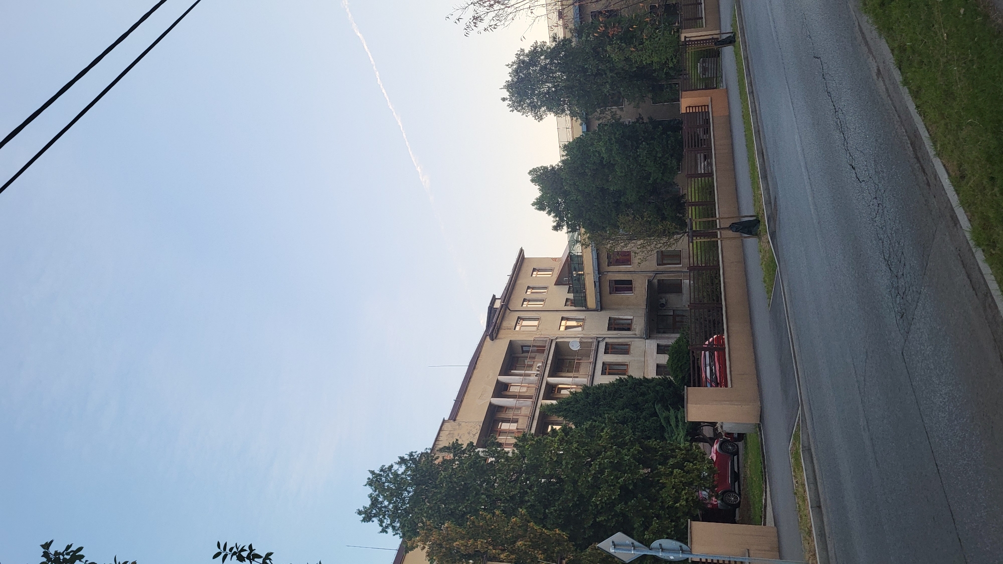 The image shows a multi-story building, likely a hospital or institutional facility, with a pale yellow facade and multiple balconies. It is located in Prešov, Slovakia, as evidenced by the visual cues consistent with the architecture and urban planning typical of the city. The building is surrounded by trees and a paved road with a sidewalk and a low wall. Two red cars are parked on the side of the road in front of the building. The sky is clear with a faint contrail. The overall scene suggests a peaceful morning or late afternoon in a suburban or urban setting.