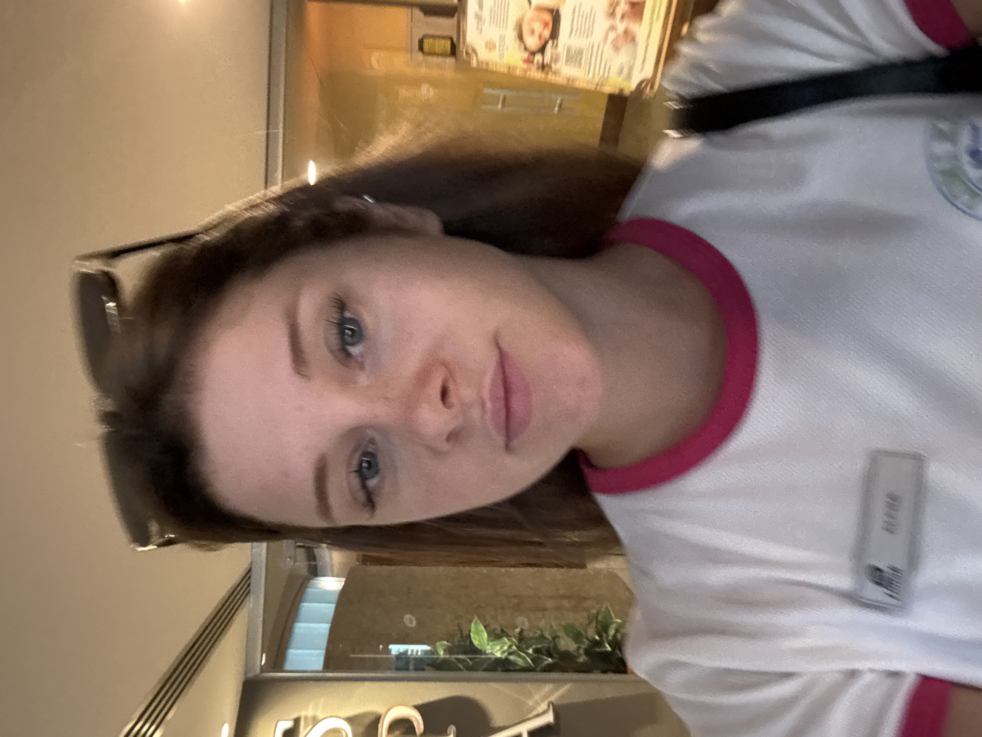 The image shows a close-up selfie of a young woman with light skin and blue eyes, wearing sunglasses on top of her head and a white t-shirt with pink trim. She has long brown hair and light makeup, with visible eyelashes. A name tag is pinned to her shirt, but the text is blurred. In the background, a sign with the letter "S" is visible on the left, and a menu or information board with images of people is on the right. The environment appears to be indoors, possibly a spa or wellness center, given the presence of plants and the type of signage. The lighting is warm. The location is stated to be the city of Aksu, Turkey.