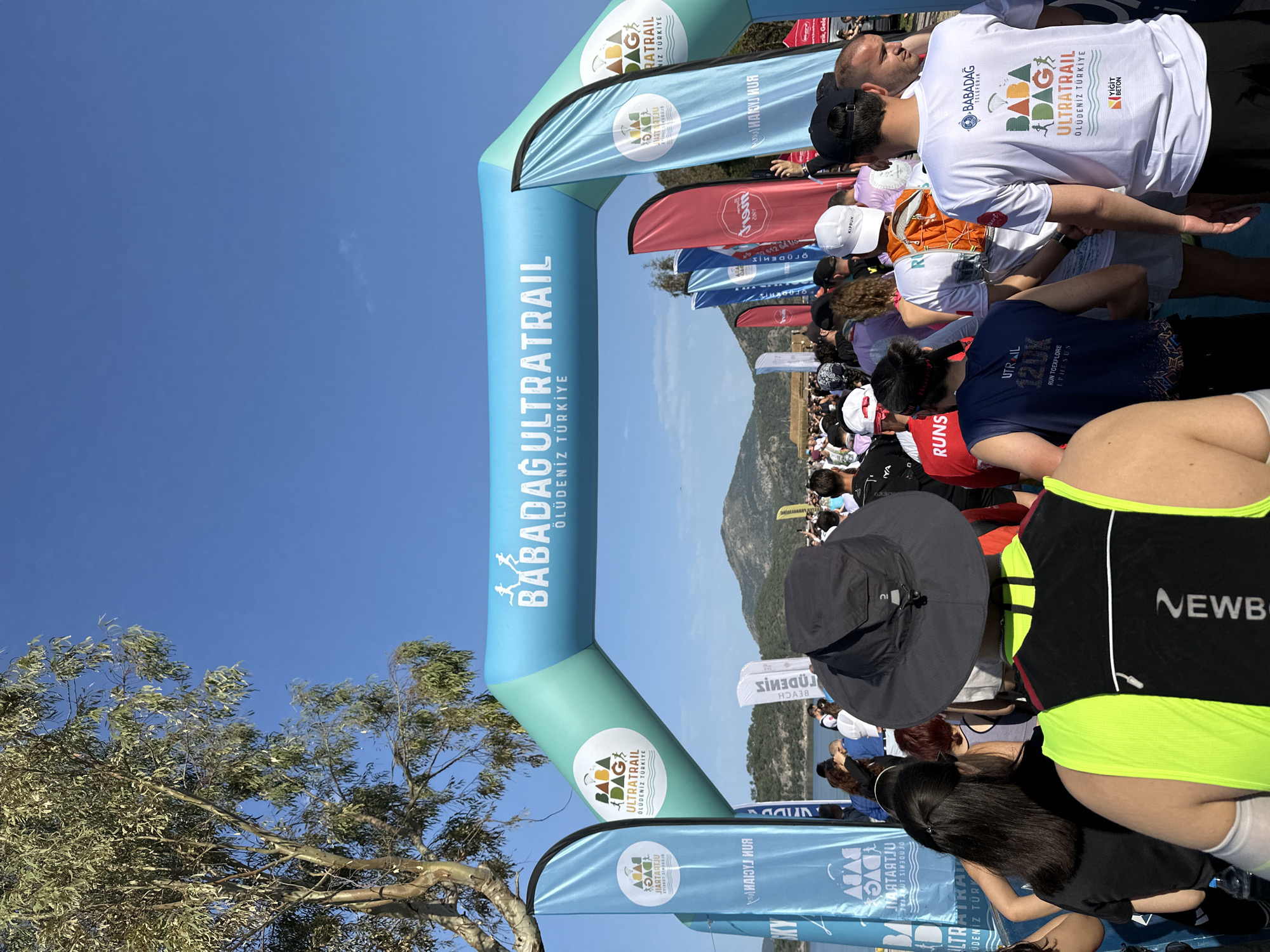 Here is a description of the image:
The image captures the starting line of the Babadağ Ultratrail race in Ölüdeniz, Fethiye, Turkey. The event is marked by a large, inflatable arch bearing the race name. Runners are gathered, preparing to begin the race.
A diverse group of people is present, indicating participants and possibly spectators. The environment features a clear blue sky. Flags and banners with sponsors' logos add to the race atmosphere. In the background, a mountain provides a scenic backdrop, representative of the terrain involved in the Ultratrail.