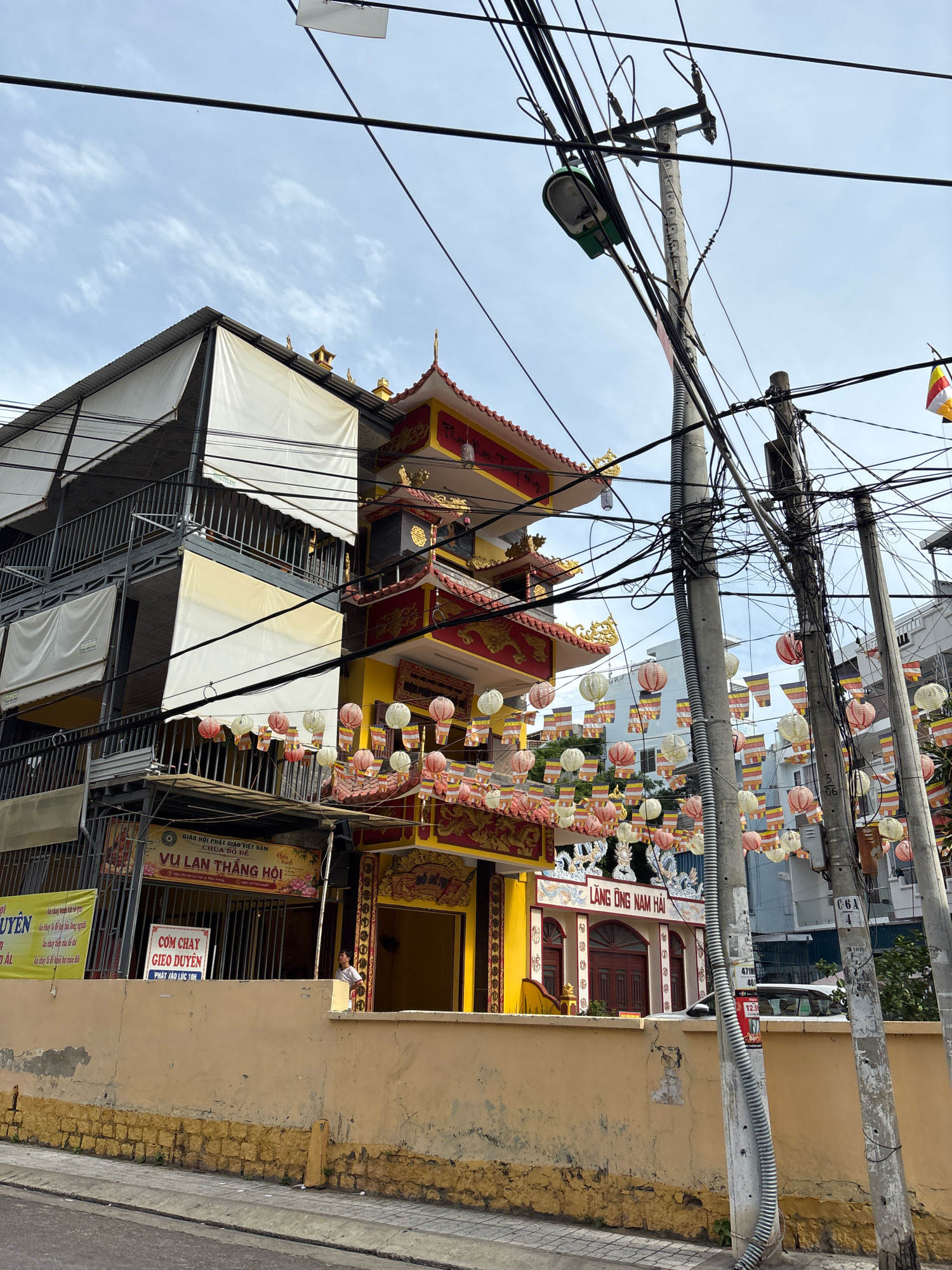This image depicts a street scene in Nha Trang, Vietnam, focusing on a building with East Asian architectural elements, likely a temple or pagoda. The building is brightly colored, with a yellow facade and red and gold ornamentation on its tiered roof. Numerous strings of colorful paper lanterns and prayer flags are suspended across the street and in front of the building, suggesting a festive or religious occasion.
To the left of the temple is a multi-story building covered in white awnings, obscuring its details but appearing to be a more modern structure. The foreground shows a paved street sloping upwards, with a yellow concrete wall separating the street from the building complex. Several utility poles with a dense network of electrical wires dominate the right side of the frame, indicating an urban environment.
Signs in Vietnamese are visible on the buildings. One sign above the temple entrance reads "LÀNG ÔNG NAM HẢI". Other signs on the left building include "VU LAN THANG HOI" and "CƠM CHAY GIO DUYEN". A person in a light-colored shirt is visible standing near the entrance of the temple. The sky is cloudy.