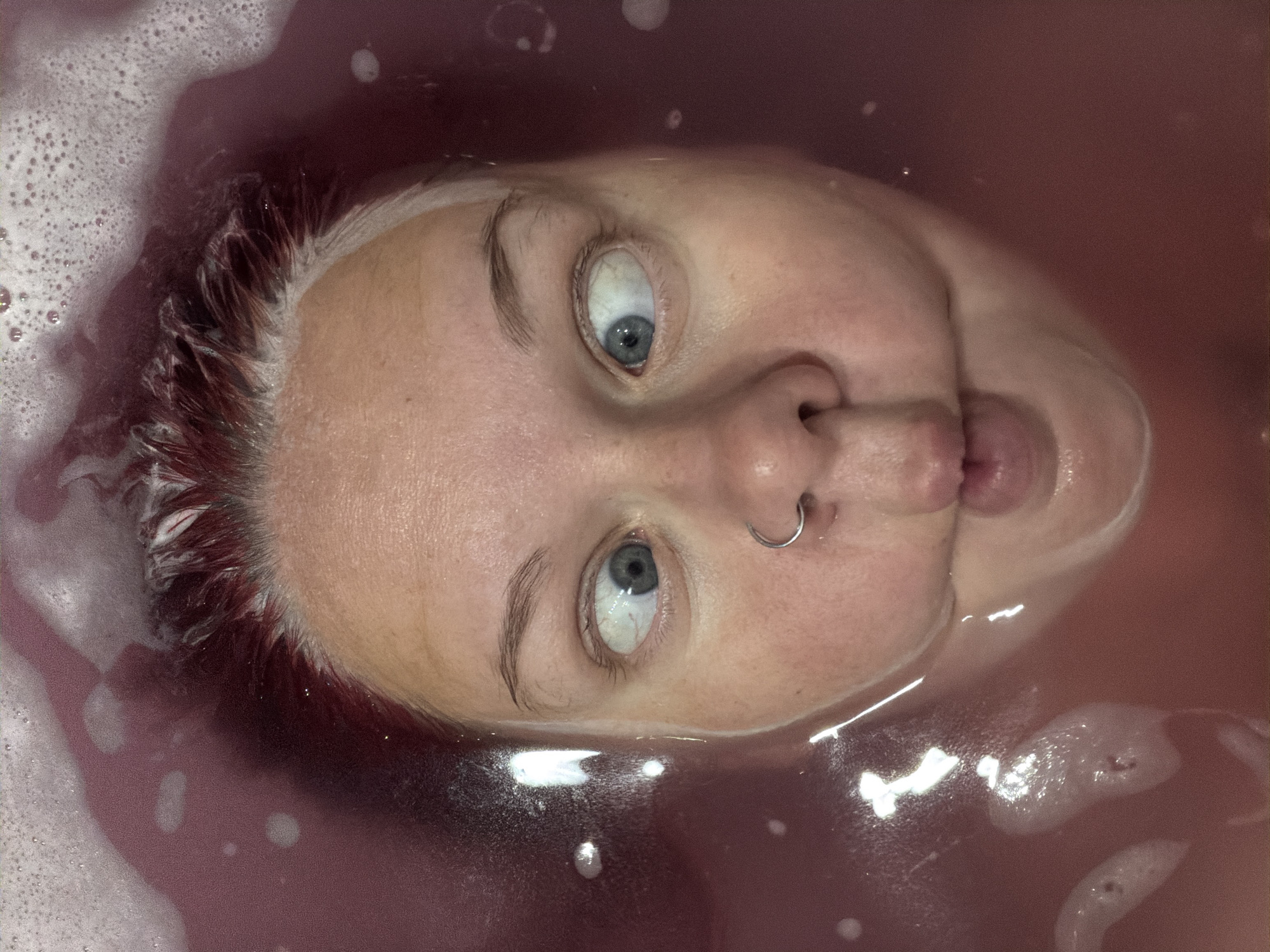 A close-up overhead view of a person's face submerged in reddish-purple bathwater. The person's eyes are crossed, and their mouth is puckered as if blowing a bubble. Red-dyed hair is visible at the top of the frame, with white foam from bath products surrounding it. A silver hoop nose ring is visible on the person's right nostril. The water is slightly murky and contains some small bubbles. There is no indication of the city of Chaikovsky, Russia, in the image.