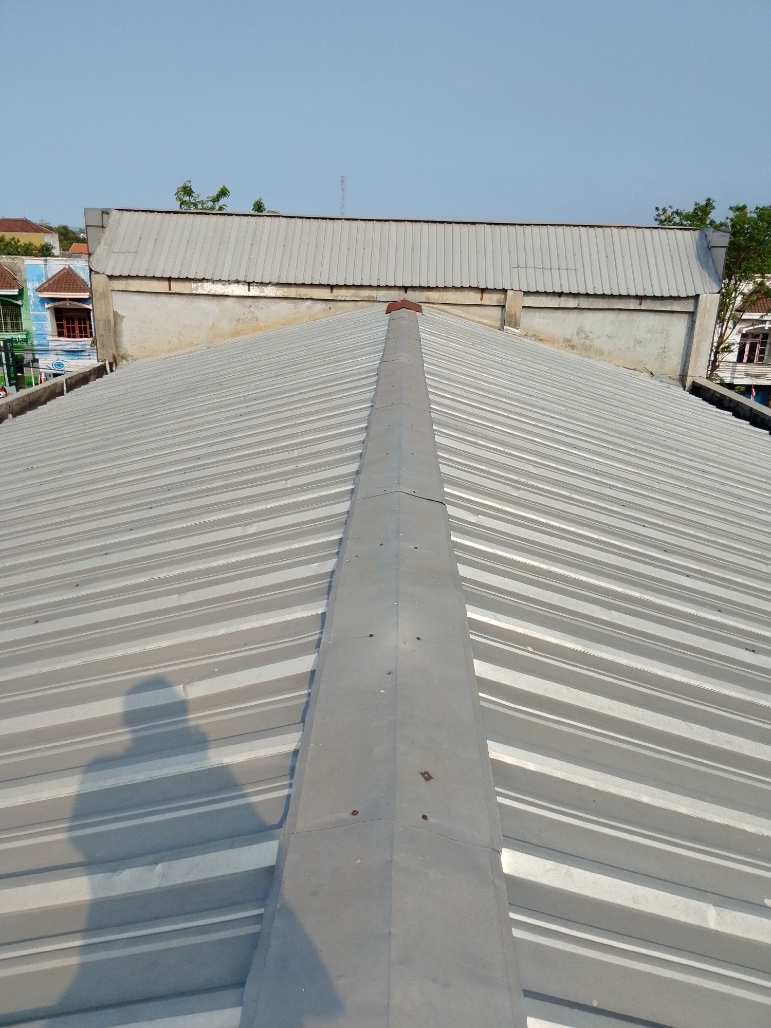 The image shows a view from a rooftop in Pasuruan, Indonesia, looking towards a long, corrugated metal roof with a central ridge cap. The roof is light gray and shows the parallel lines of the corrugated material. The shadow of a person is cast on the left side of the roof. In the background, there are other buildings with various roof types, including what appears to be a tiled roof and another corrugated metal roof with a different profile. Trees and a clear blue sky are visible above the buildings. The architectural style suggests an urban or semi-urban environment.