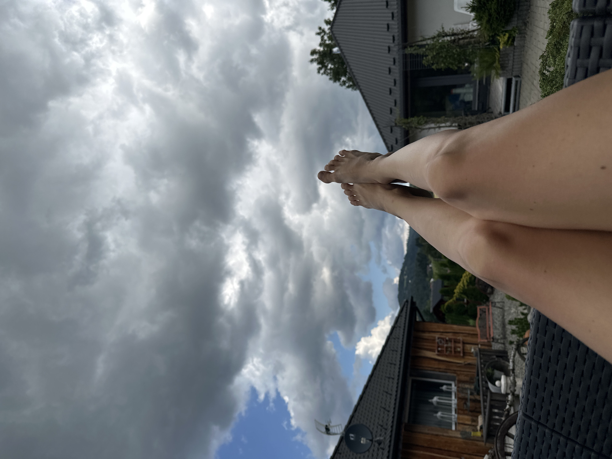 The image shows a close-up view of a person's legs and feet, positioned upwards against a backdrop of a cloudy sky and residential buildings. The legs are bare and tanned, with toes visible. The background features two houses with dark tiled roofs. One house on the left has a wooden facade, while the house on the right appears more modern. The sky is filled with heavy, dark clouds, suggesting overcast weather, with patches of blue sky visible. The foreground is occupied by a dark, woven-textured surface, possibly a piece of furniture. The scene is set outdoors, likely in a backyard or patio area of a home in Wisła, Poland.