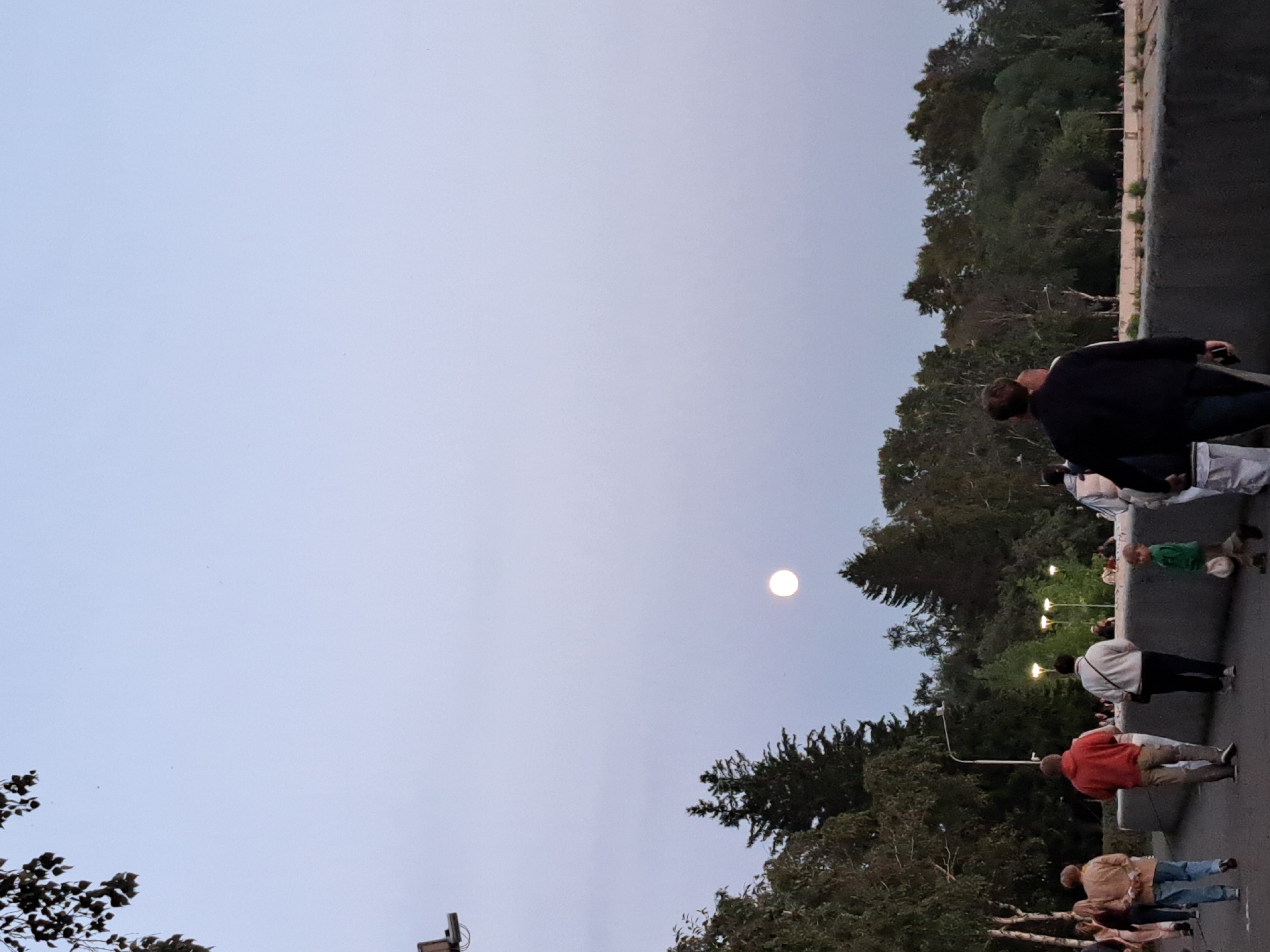 The image depicts an outdoor scene, likely in a park or along a waterfront, in Irkutsk, Russia, during twilight. The sky is a pale, dusky blue with a full moon visible. In the foreground, a group of people are walking away from the camera on a paved path. Among them are a woman in a beige jacket and blue jeans, a man in a red hoodie and beige pants, and a woman in a white sweater and dark pants. A young child in a green shirt is also present, holding a hat. Behind them, a low, grey concrete barrier runs horizontally, separating the path from a wooded area with numerous trees and some lampposts. The trees are dense and form a dark backdrop against the sky. The overall impression is one of a leisurely evening stroll in a city park.