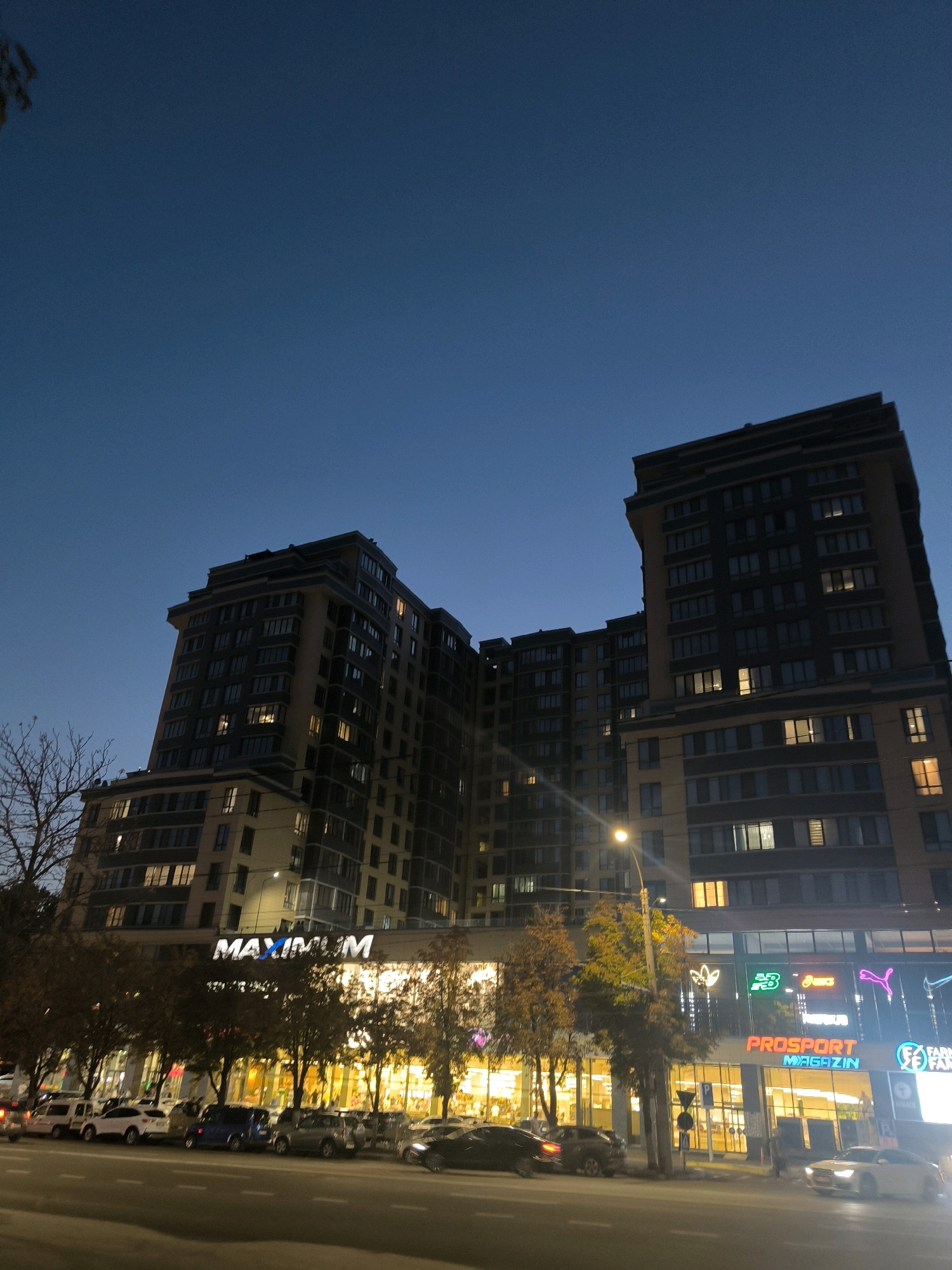 The image shows a wide shot of a modern apartment building with commercial spaces on the ground floor, captured at dusk in Chișinău, Moldova. The building has a distinctive architectural style with multiple setbacks and a dark facade contrasting with illuminated windows. The commercial spaces feature prominent signage for "MAXIMUM" and "PROSPORT MAGAZIN," along with logos for brands like Adidas, New Balance, and Asics, indicating a retail and entertainment hub. Several cars are parked along the street in front of the building, and a few trees with autumn foliage are visible. The sky is a deep twilight blue, suggesting the time of day. A streetlamp is also visible, emitting a warm glow.