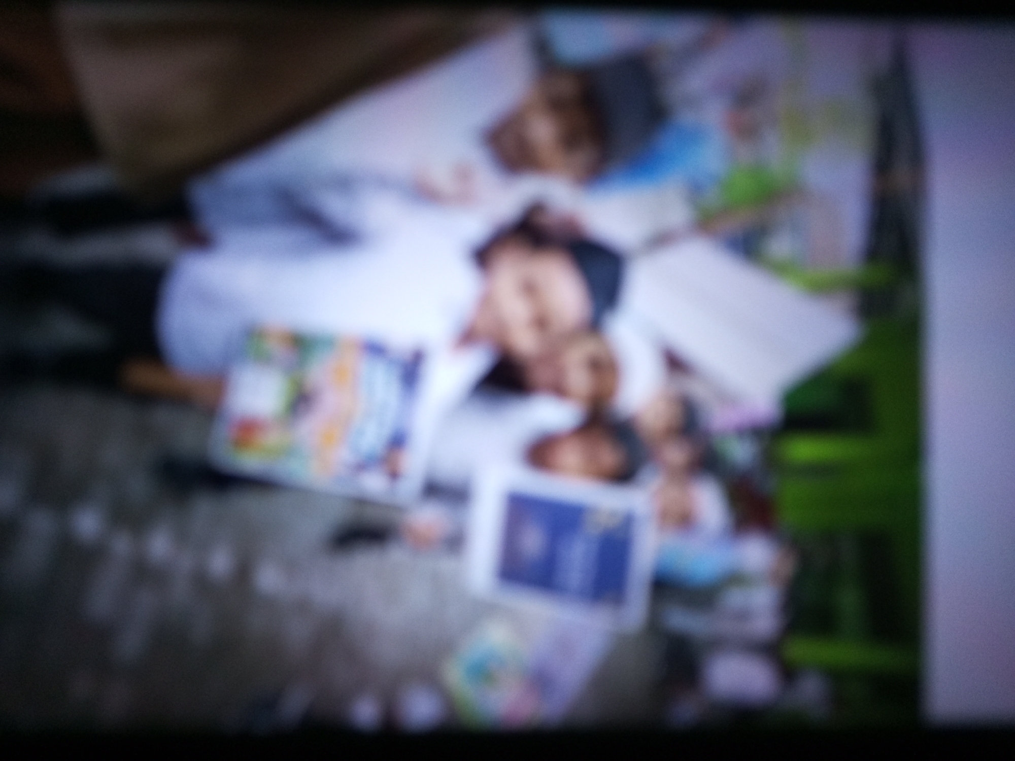 The image depicts a blurry, low-resolution scene showing several people, likely children, lying down on a floor or ground. One individual in the foreground is wearing a white shirt. There are several colorful objects scattered around, resembling books or toys. The background is indistinct, with hints of green and dark areas, suggesting an outdoor or semi-outdoor setting. The image appears to be taken from a distance and is heavily pixelated, making precise identification of details challenging. The scene does not provide any specific information that could be used to determine the location in Banyudono, Indonesia.
