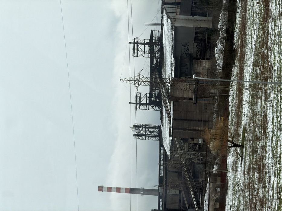 The image depicts an industrial landscape in Terjola, Georgia, under an overcast sky with patchy snow covering the ground and structures. In the foreground, a field of dry, sparse vegetation is visible, lightly dusted with snow, revealing brown earth. A single deciduous tree, bare except for a small amount of lighter foliage at its crown, stands left of center. To the far left foreground, a small, low-rise building with a dark red roof is partially visible. A grey streetlamp pole stands vertically on the right side of the foreground.
The midground and background are dominated by a large industrial complex. On the far left, a tall, cylindrical industrial chimney with alternating red and white horizontal stripes rises prominently. The complex consists of multiple dark, multi-story buildings constructed from concrete and metal, many of which have roofs and elevated platforms lightly coated with snow. Some building facades display dark rectangular openings, possibly windows, with several appearing to be broken or empty. Numerous lattice-work metal structures, including what appear to be support towers, a tall power transmission tower with multiple lines traversing the scene, and conveyor belt systems, are integrated throughout the complex. The conveyor belts, also dusted with snow, ascend and descend between various sections of the buildings. Another similar power transmission tower is visible further left within the complex. No individuals or discernable human activity are present in the scene.