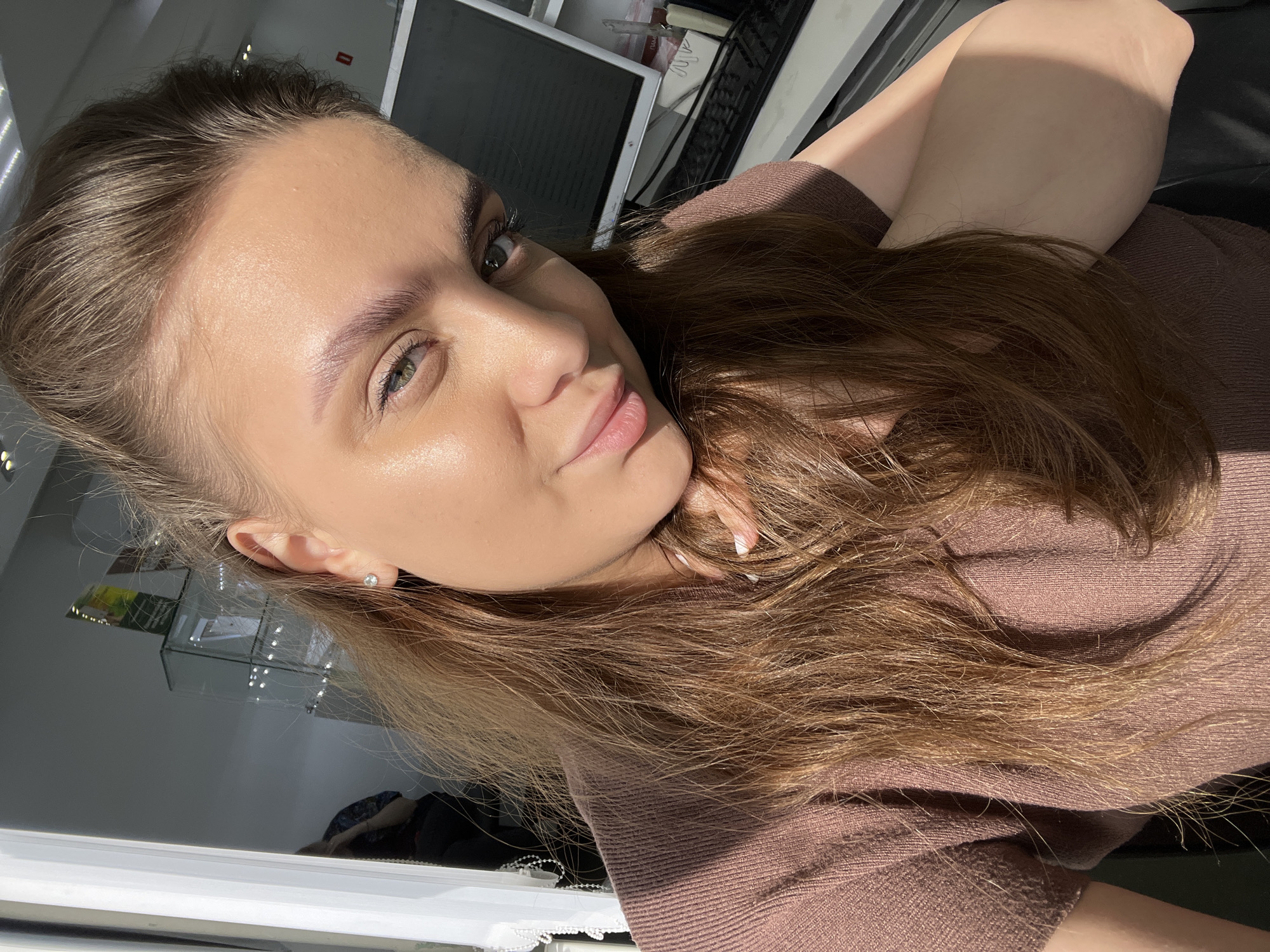 A young woman with light brown hair, styled back from her face, is posing for a selfie. She wears a light brown top and a small stud earring. Her eyes are green, and she has a light complexion. Her right hand is near her face. In the background, a monitor, keyboard, and other office items are visible. The photo appears to have been taken indoors in a brightly lit room. The exact location in Chaikovsky, Russia, is impossible to determine from the image.