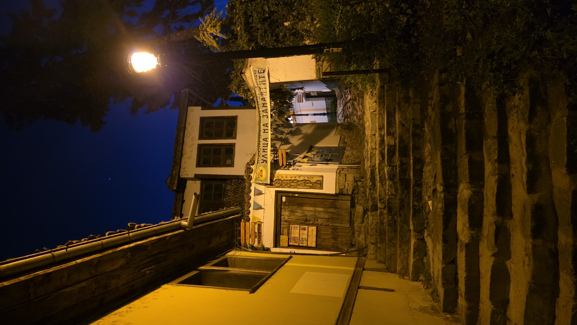 A street scene at night in Plovdiv, Bulgaria, shows a narrow stone staircase leading uphill between buildings. A street lamp illuminates the scene from the left, casting a warm glow. On the left, there is a building with a white facade and dark-framed windows. Above a wooden door, a sign in Cyrillic reads "Улица на занаятите" (Street of Crafts). A second building is visible in the foreground, also white with a dark roof. The right side of the staircase is lined with a stone wall overgrown with vegetation. The sky is dark blue, and a tree is partially visible at the top of the frame.
