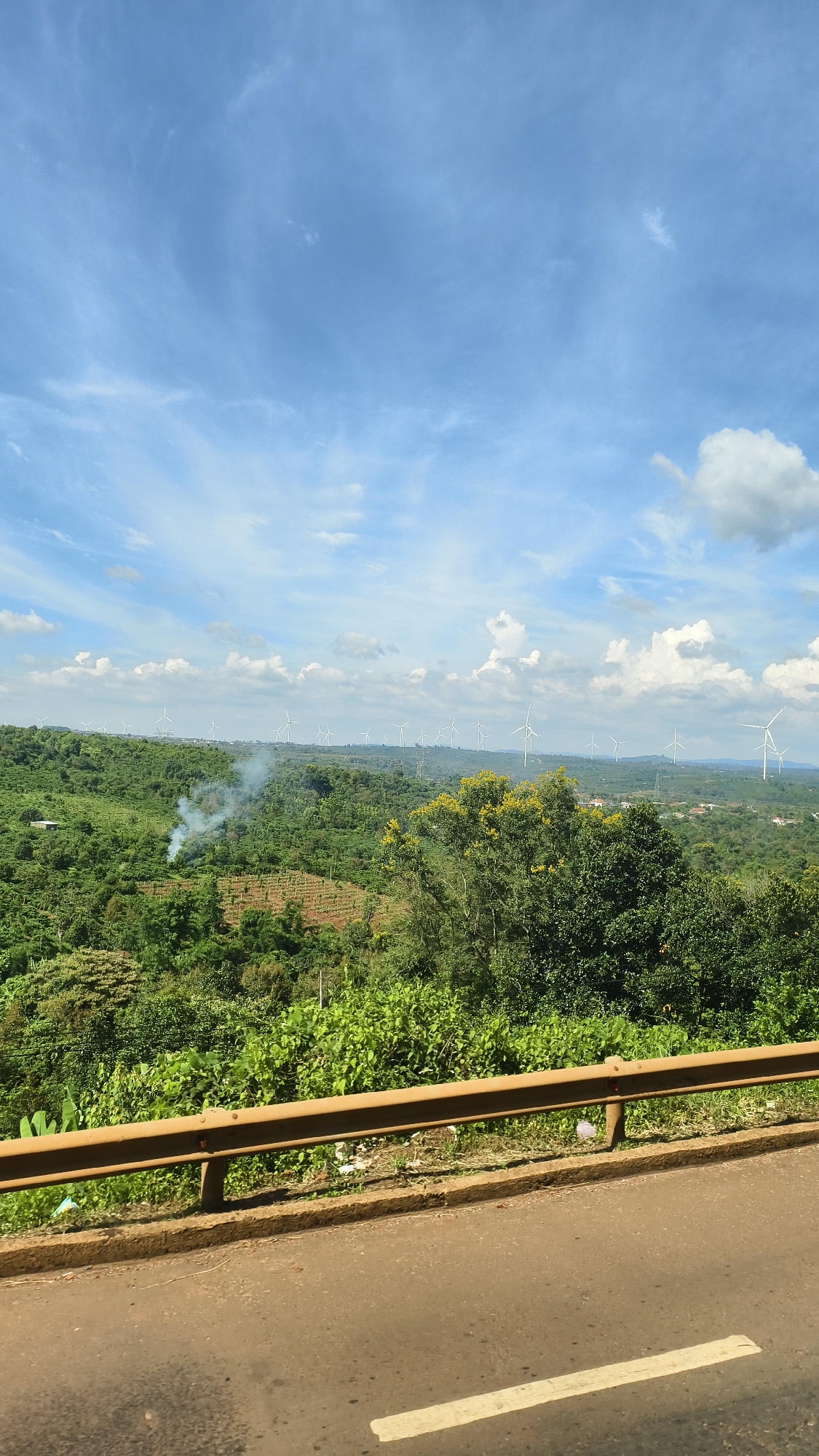 A wide-angle, outdoor, eye-level shot captures a lush green landscape under a partly cloudy blue sky. In the foreground, a dirty brown guardrail separates a two-lane asphalt road from the elevated terrain. The road has a faded white line marking the lane. The terrain is densely covered with various shades of green foliage, including trees and smaller bushes, with some patches of exposed reddish-brown soil, possibly indicating agricultural activity. In the mid-ground, a small plume of white smoke rises from the vegetation. Scattered across the horizon are numerous white wind turbines, suggesting a wind farm. The sky is a vibrant blue with scattered white clouds, some of which are wispy and others more puffy. The overall scene conveys a sense of natural beauty and renewable energy infrastructure.